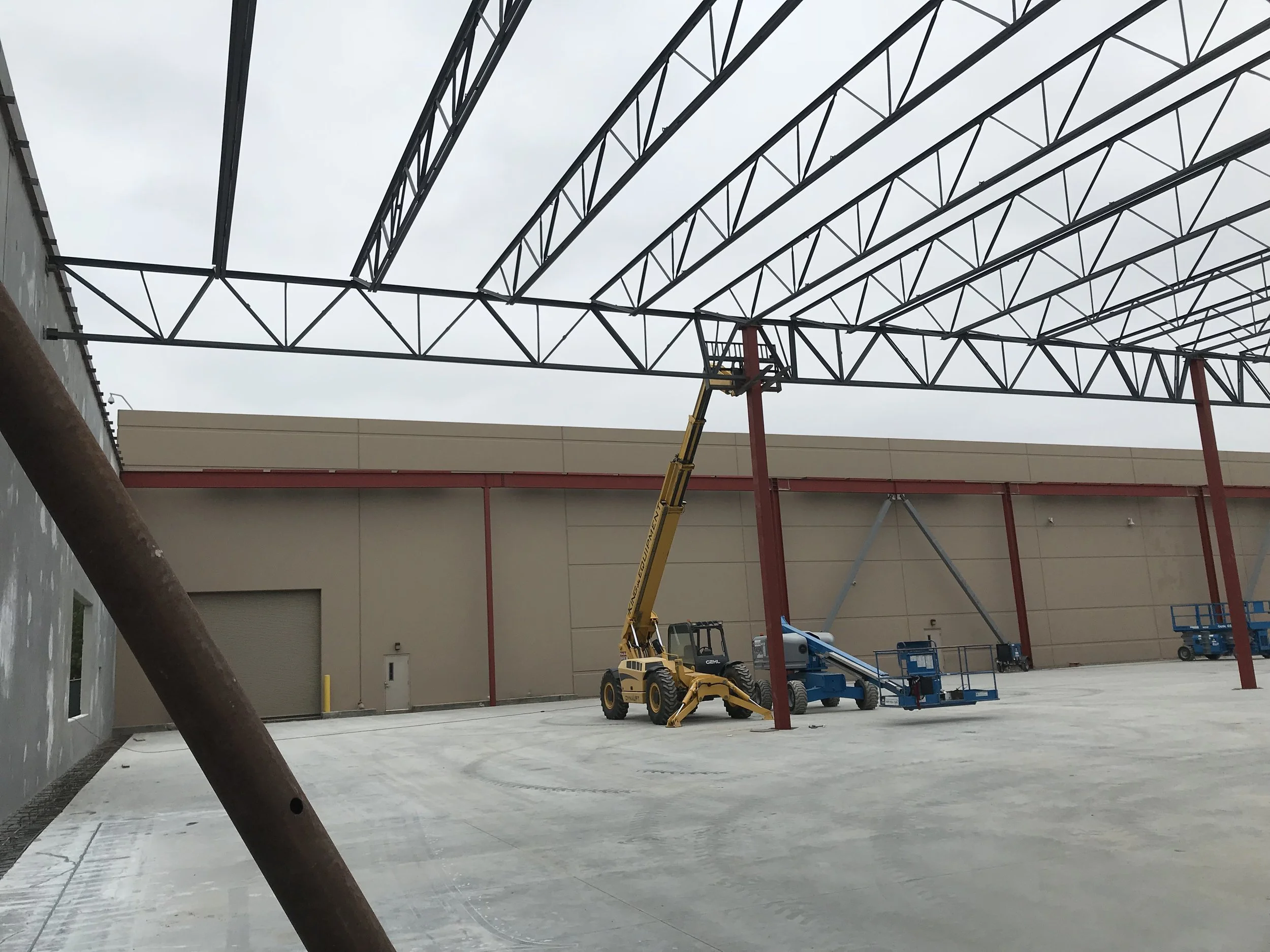 Construction site with a yellow boom lift and blue scissor lift, steel framing for a building roof, and cloudy sky in the background.