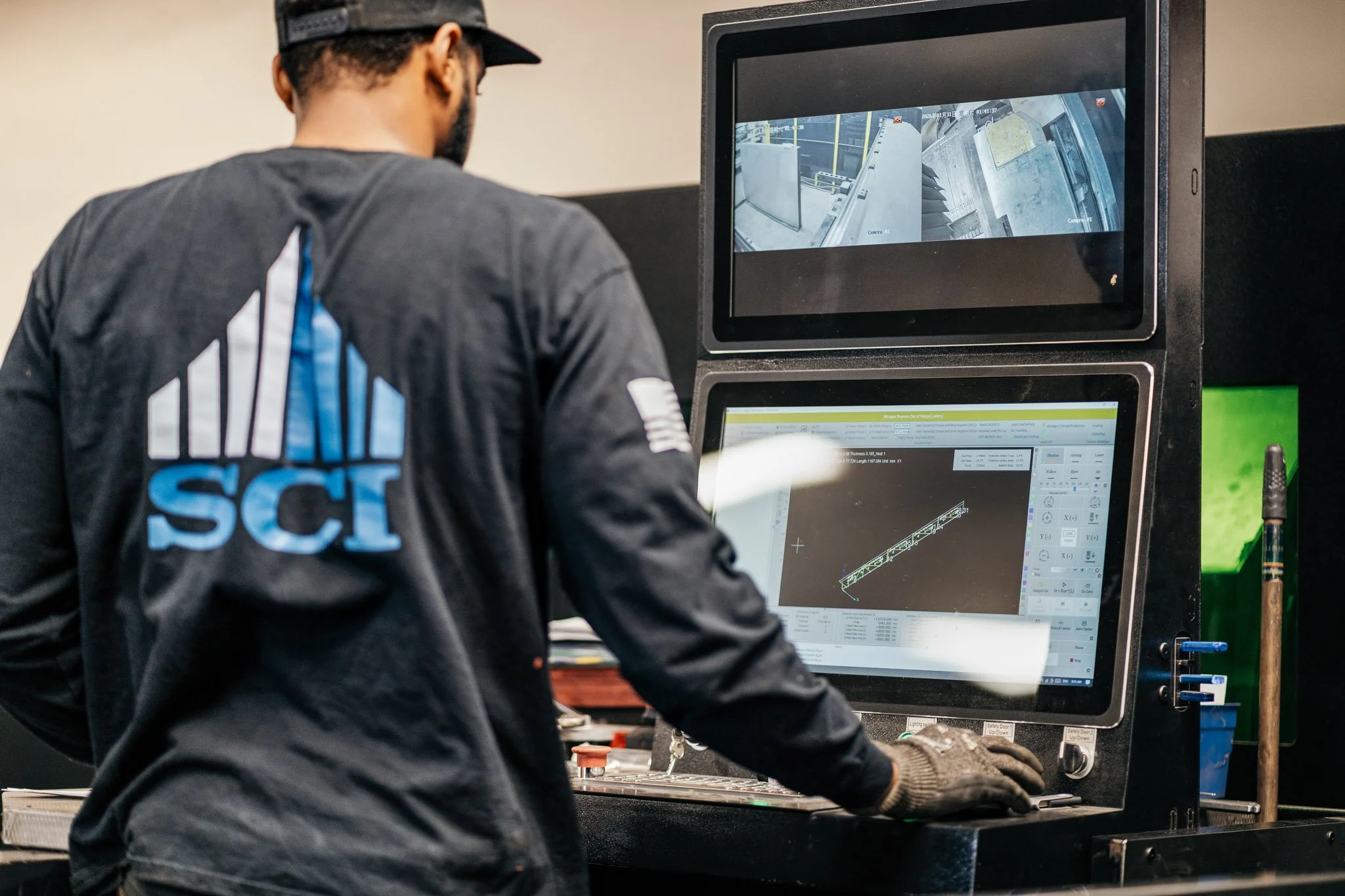 A man wearing a black cap and a dark shirt working at a computer station with multiple screens, one displaying technical or engineering software and another showing a video feed from surveillance cameras, in an industrial or workshop setting.