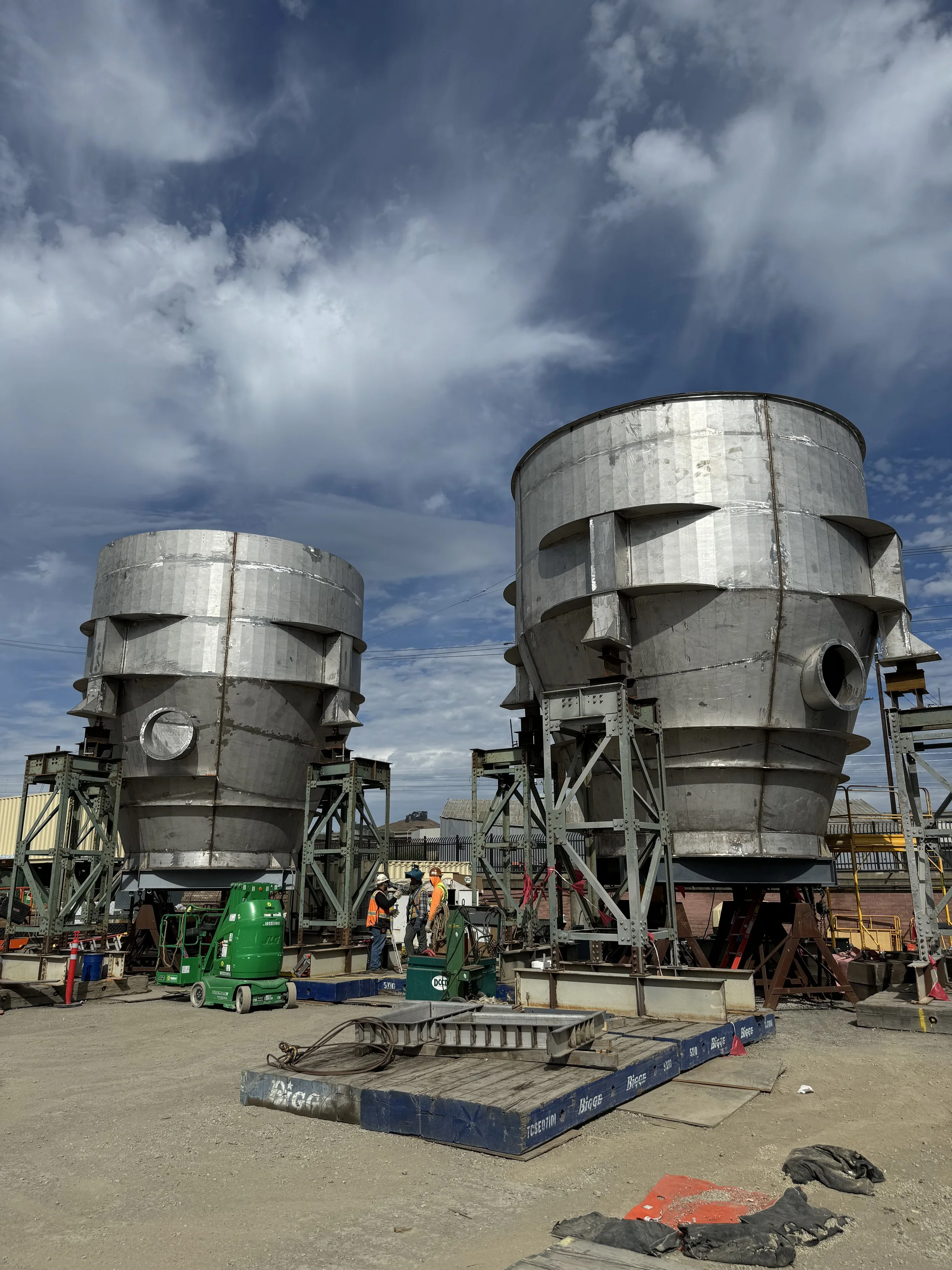 Construction site with two large, metallic, industrial structures being assembled, workers in safety gear, and construction equipment on the ground under a cloudy sky.