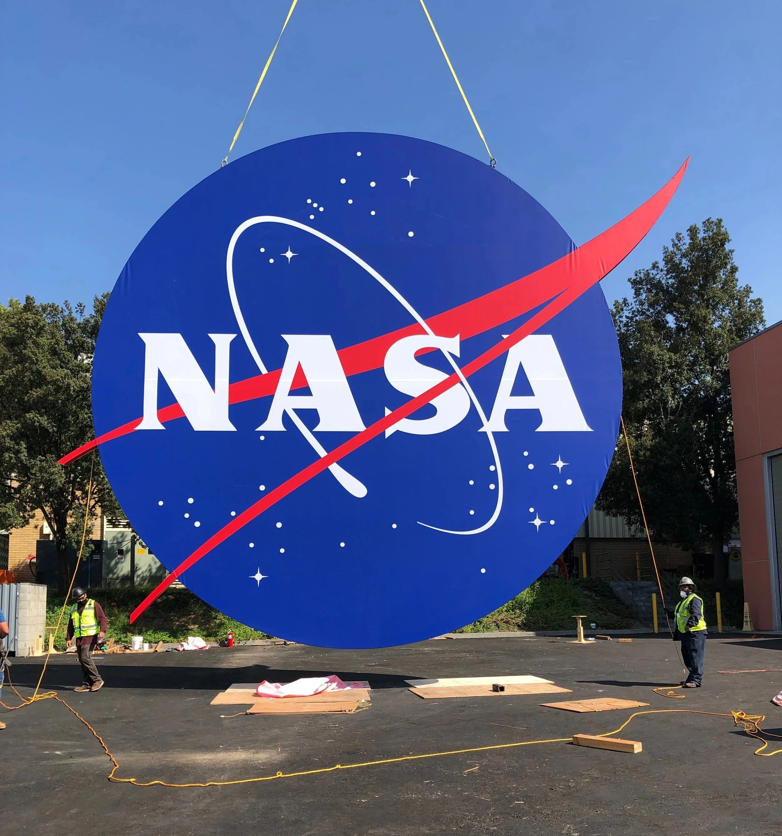 Workers preparing to hang a large NASA logo banner outdoors during daytime. The banner is blue with white stars and features the NASA logo with a red swoosh. Two workers are visible, wearing reflective vests and helmets, handling ropes and tools on a black asphalt surface with some wooden planks.