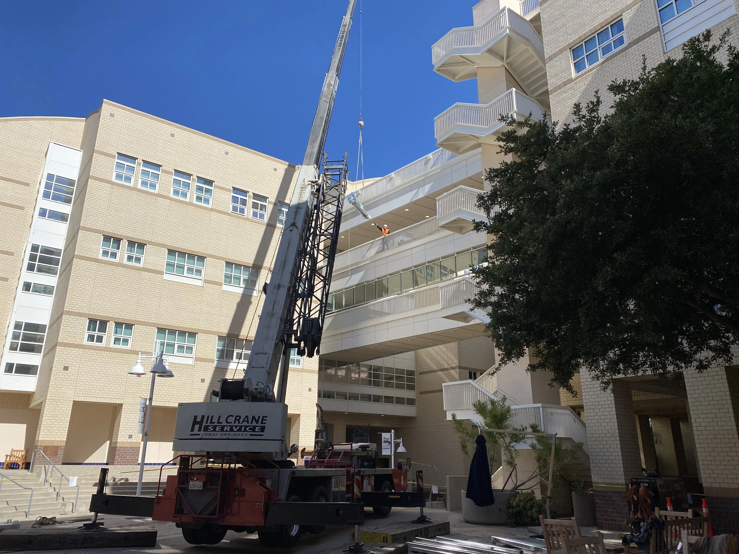 A large construction crane lifting objects in front of a modern residential building with multiple balconies, large windows, and brick exterior. Construction workers are visible on the balcony area, and the scene is set under a clear blue sky.