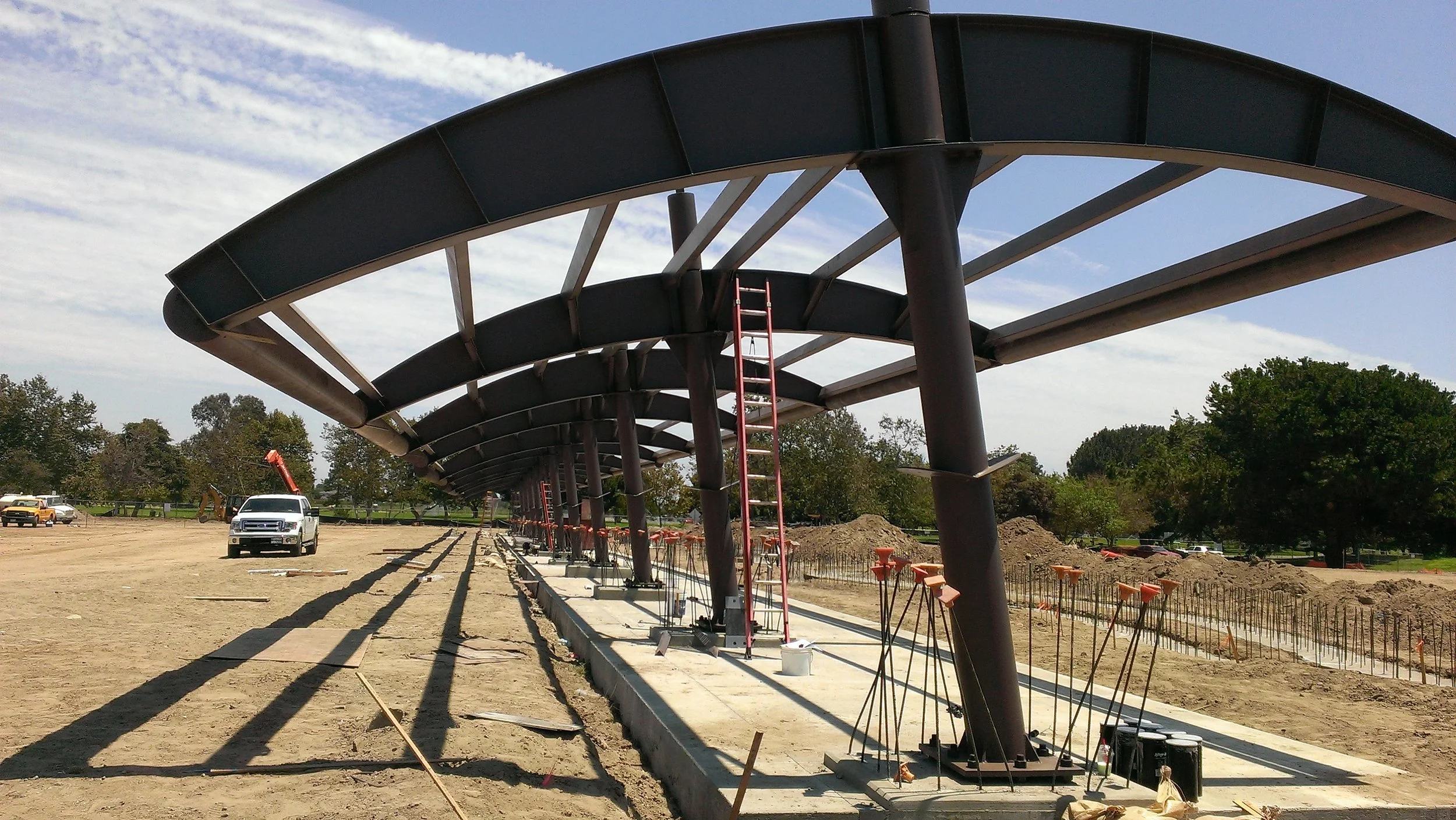Under construction of a covered walkway with large dark panels, red ladders, construction vehicles, and tools on a dirt lot with trees and a partly cloudy sky in the background.