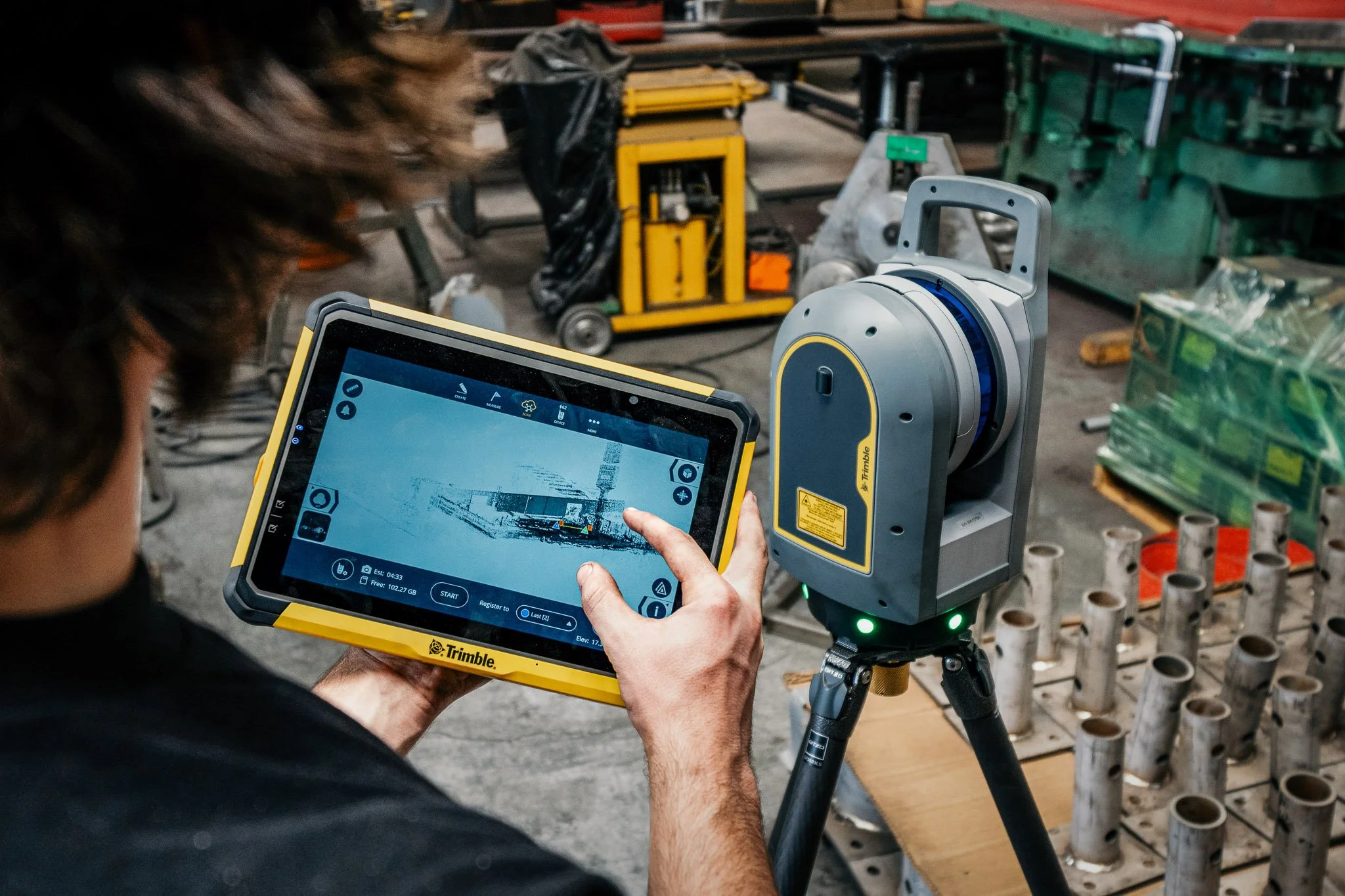 A person holding a touchscreen tablet controlling a robotic device in a workshop or industrial setting.