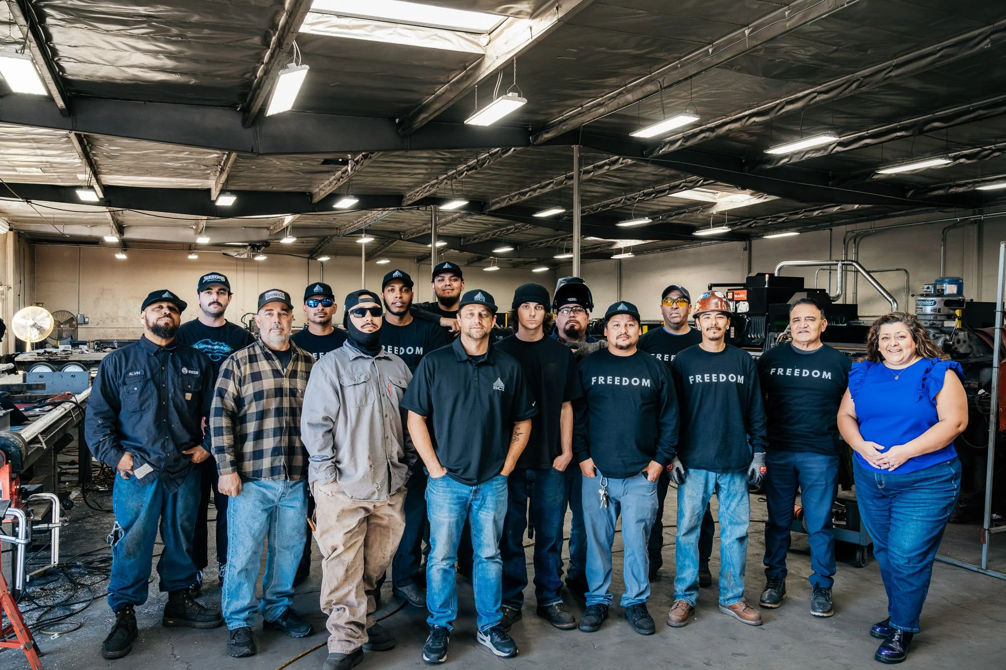 Group of diverse workers in casual and work attire standing together in an industrial warehouse or factory setting with machinery and tools in the background.