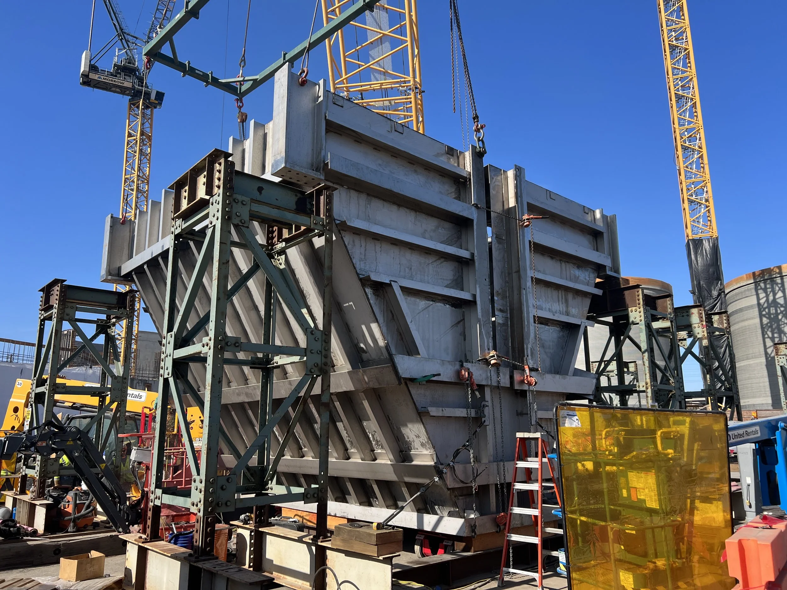 Construction site with steel framework and crane, workers' equipment, and safety barriers under a clear blue sky.