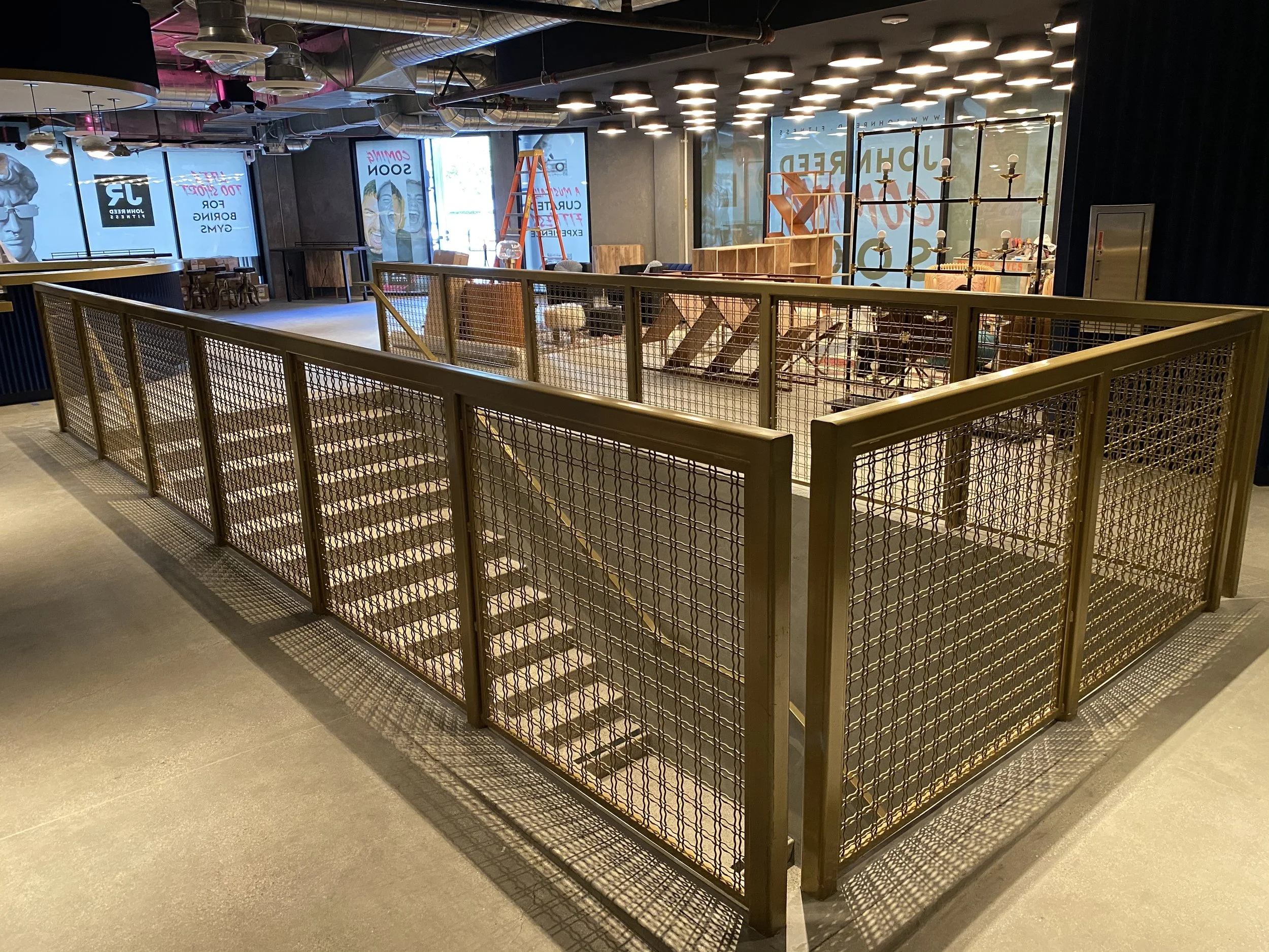 Empty indoor area with steel security fencing, overhead lighting, and construction materials including a standing orange ladder and wooden shelves, suggesting ongoing setup or renovation.
