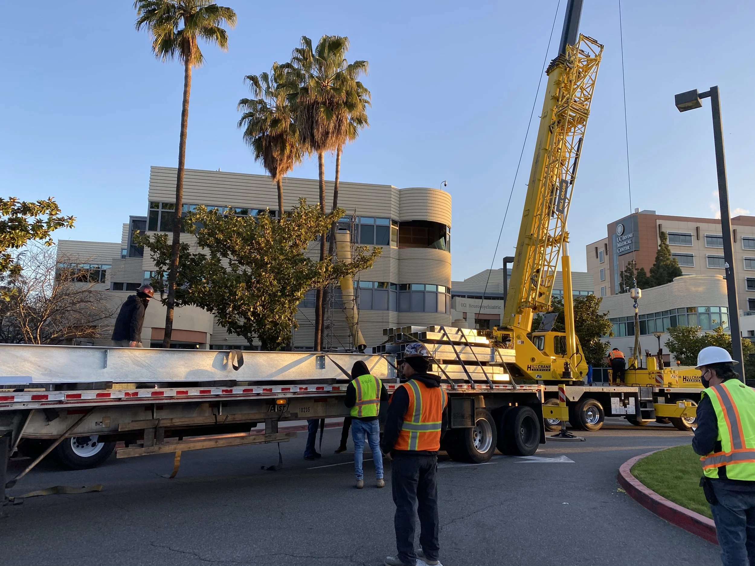 Workers in safety vests and helmets work around a large yellow crane on a flatbed truck, installing or removing a landscaping element near a building with palm trees and a medical center sign.