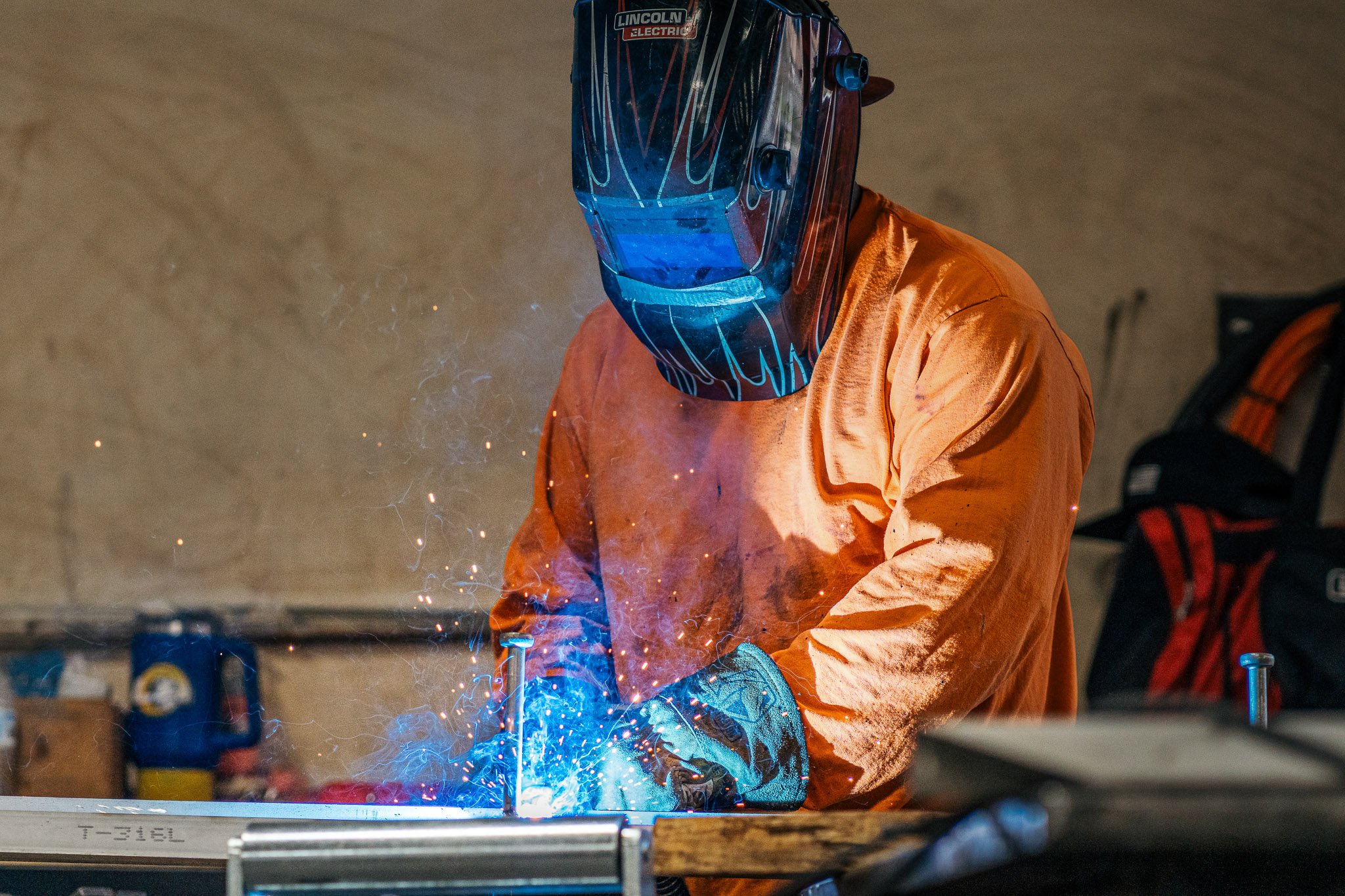 A person wearing an orange protective suit and a welding helmet with a blue tint, welding metal on a workbench. Sparks and bright blue light are visible from the welding process.