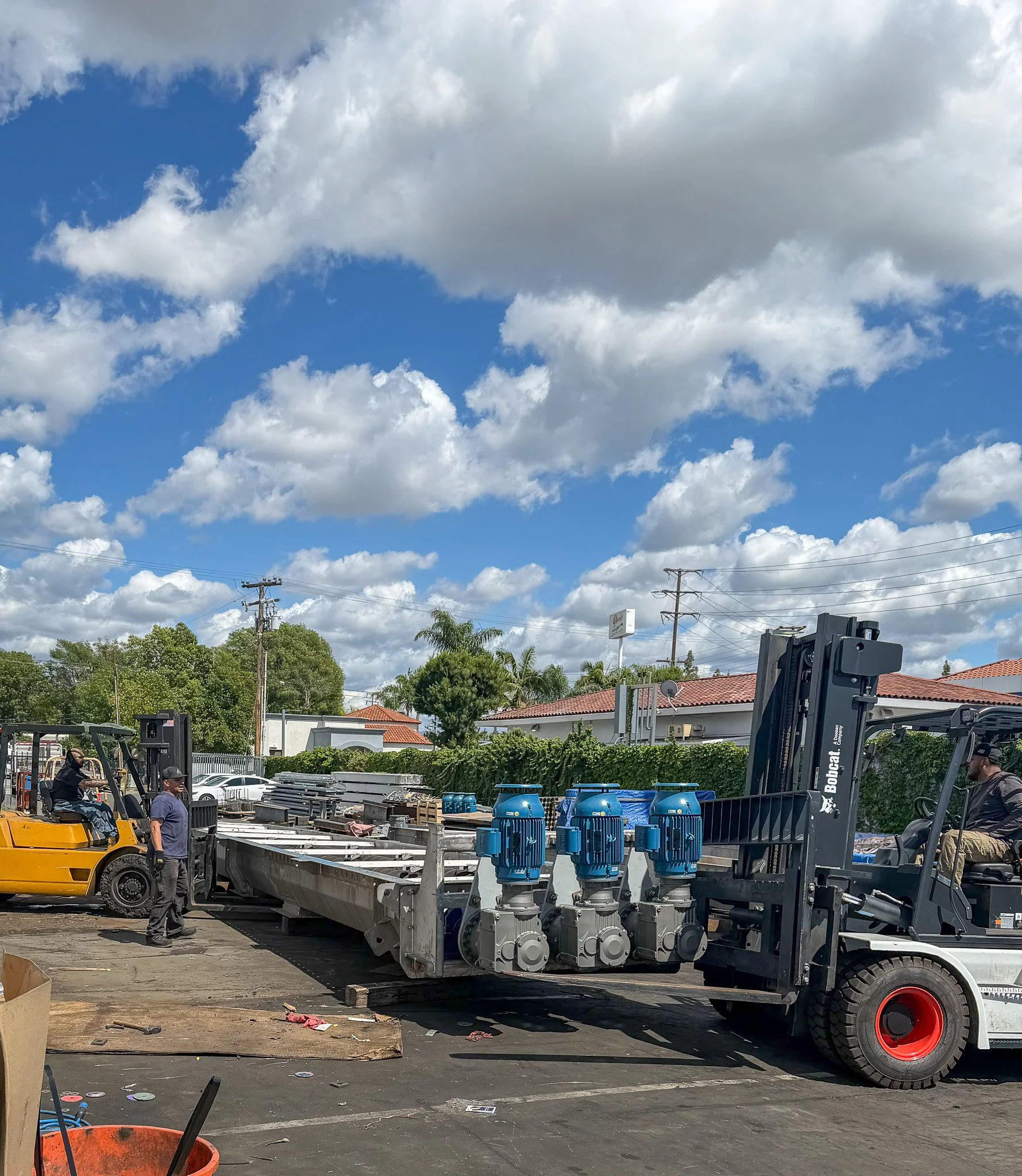 Workers in a construction or industrial setting loading large industrial equipment onto a flatbed truck with a forklift. There are blue water pressure tanks on the truck, and the background features trees, power lines, and a partly cloudy sky.