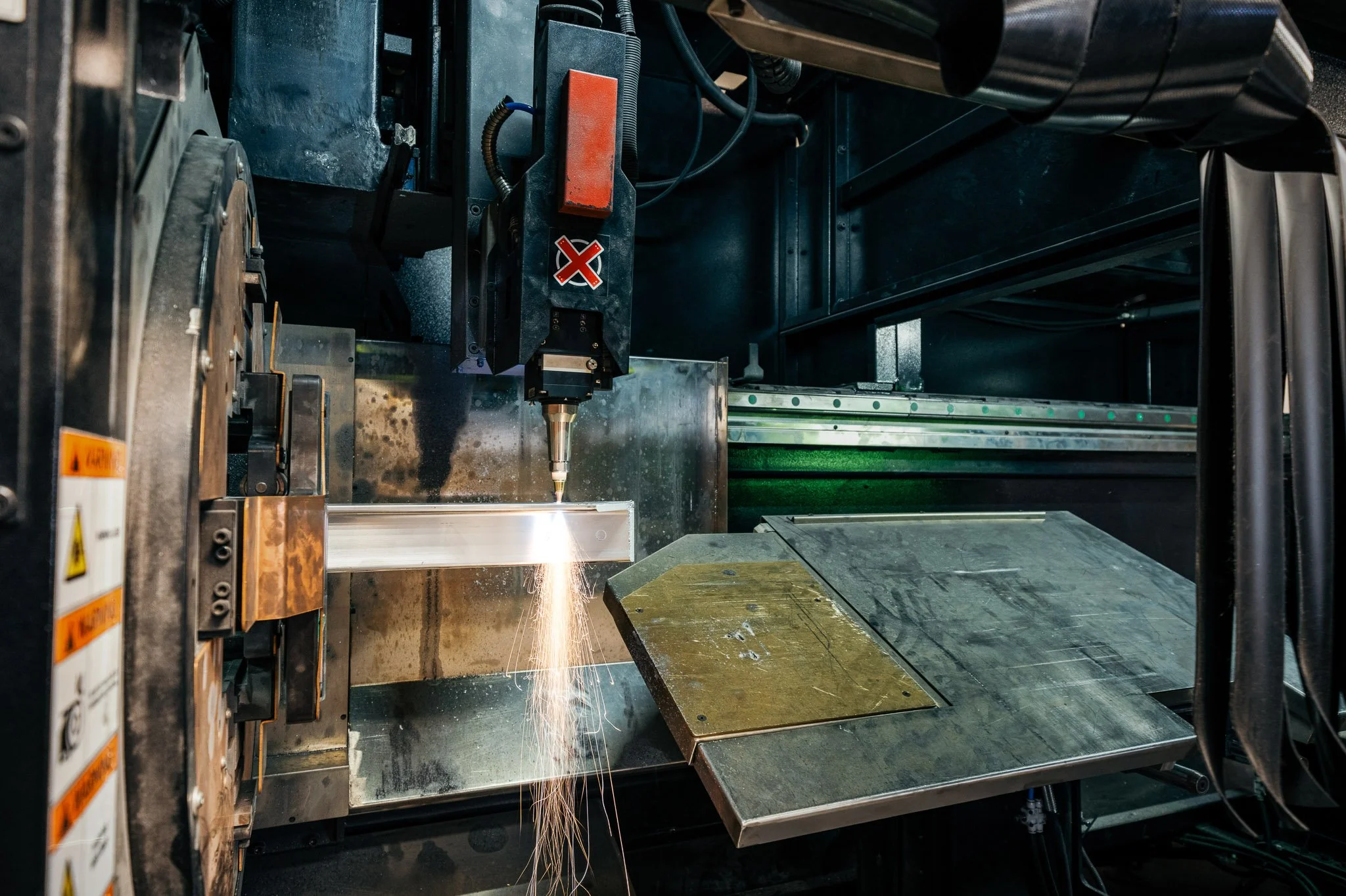 Industrial manufacturing setting with a laser cutting machine in operation, emitting sparks onto a metal sheet, surrounded by machinery and equipment.