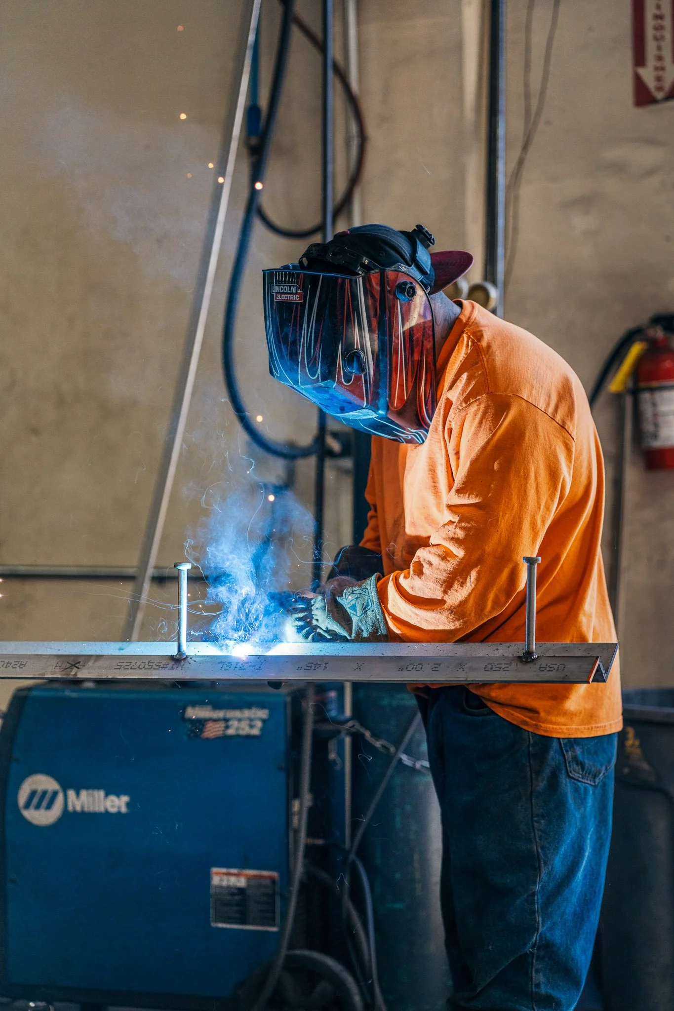 A person welding metal in an industrial workshop, wearing an orange shirt and protective helmet with a face shield, sparks and smoke visible.
