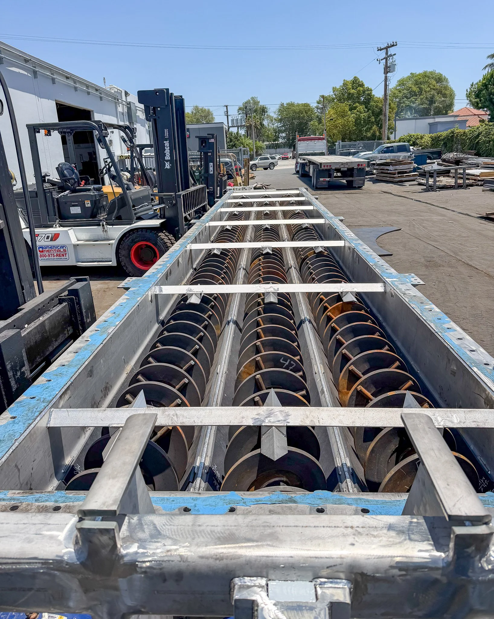 View of industrial equipment and construction materials outdoors on a sunny day, including large metal pipes, flatbed trucks, and pallets of materials near a building and power lines.