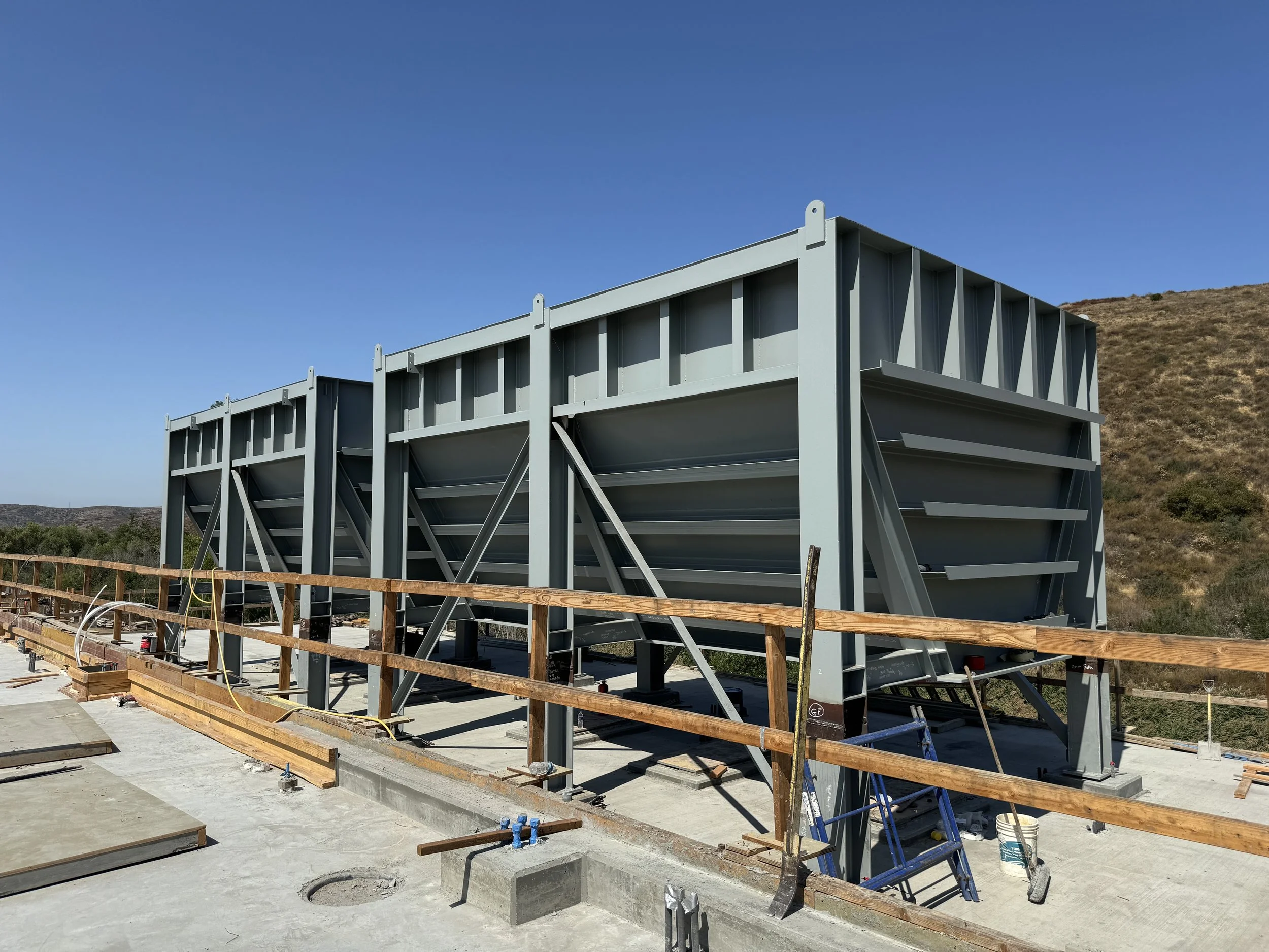 Construction site with large steel structure framework and wooden safety railing, set against a hillside and clear blue sky.