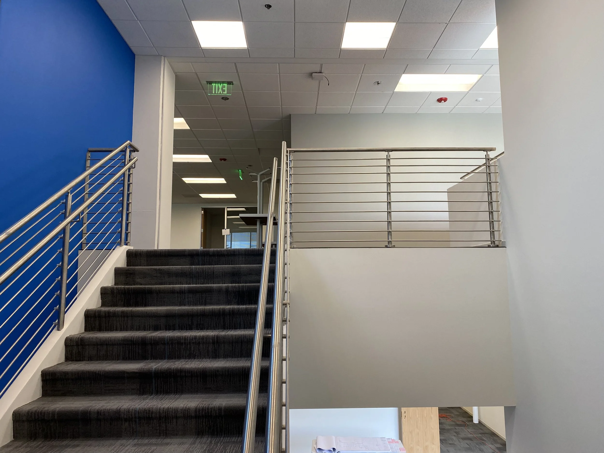 Staircase with black carpet running up to a second level, with stainless steel handrails on both sides. The staircase is located next to a blue wall, and the area has a drop ceiling with fluorescent lights. There are two green exit signs on the ceiling, and a white wall visible at the top of the stairs. The bottom of the stairs shows a partially visible open area with gray and black flooring.