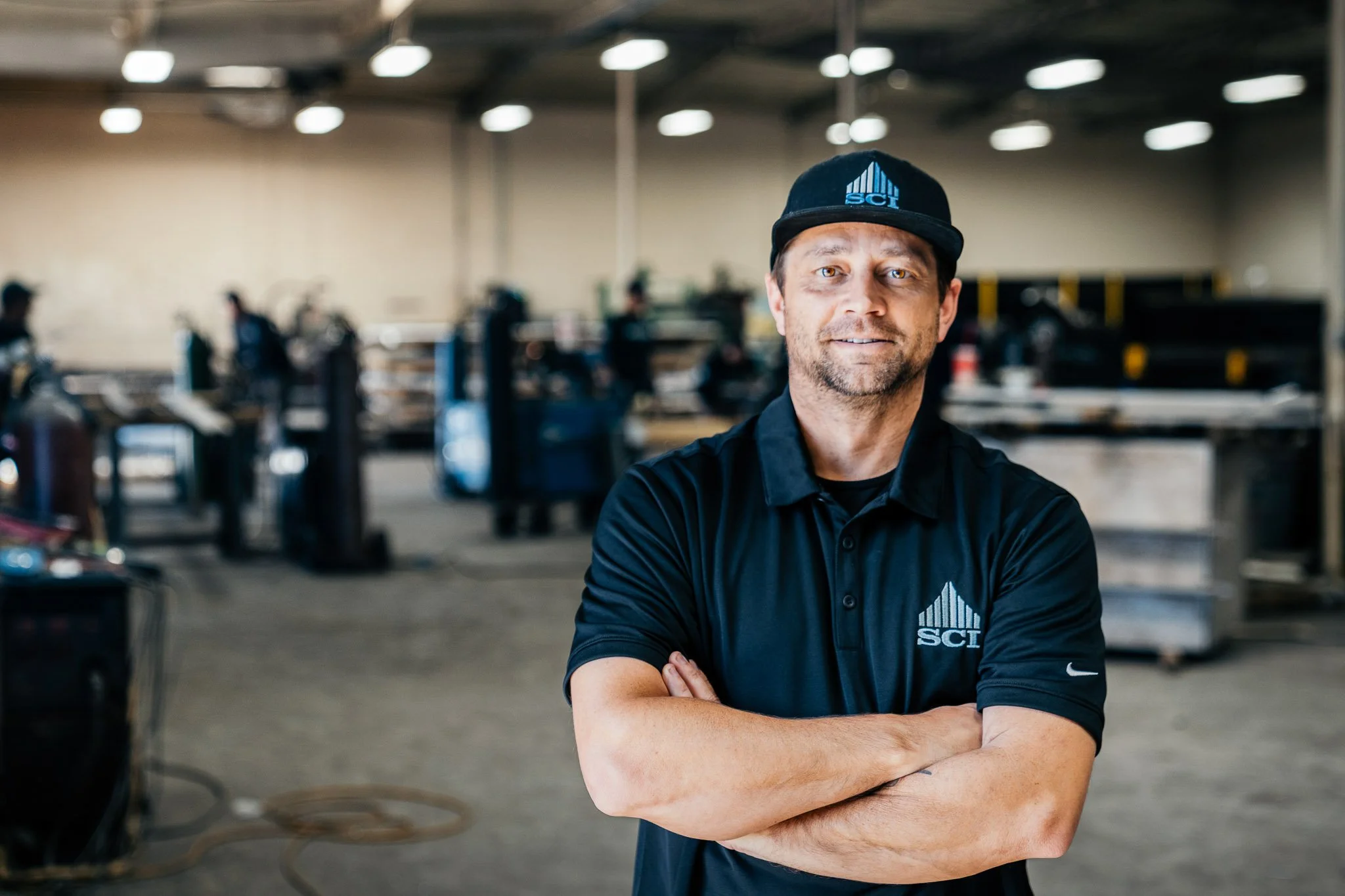 A man with brown hair and a beard wearing a black baseball cap with a blue logo and a black polo shirt with a matching logo, standing in a workshop or factory with blurred machinery and other workers in the background