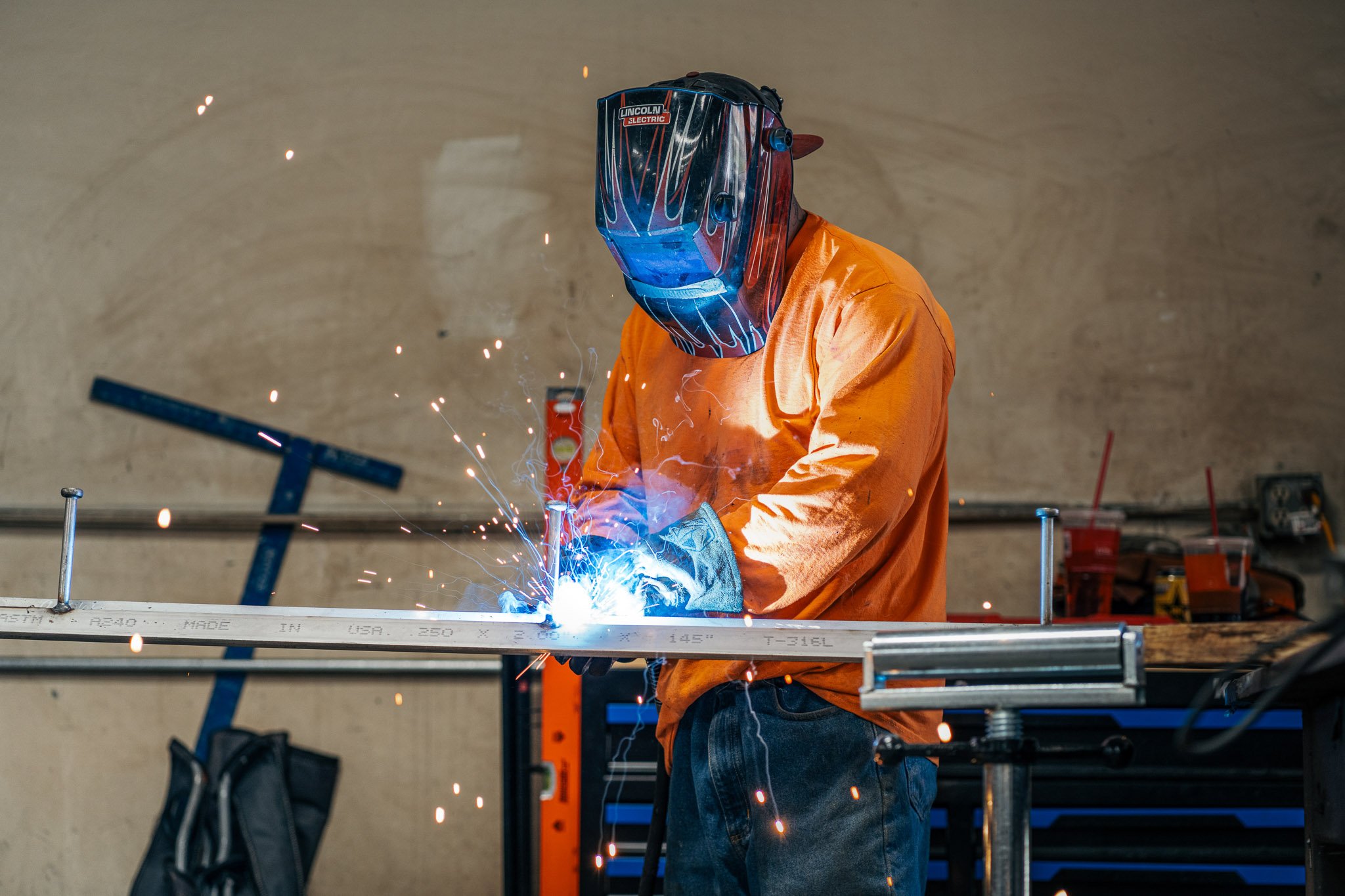 Worker welding metal in a workshop, wearing a protective helmet and orange shirt, sparks flying from the welding process.