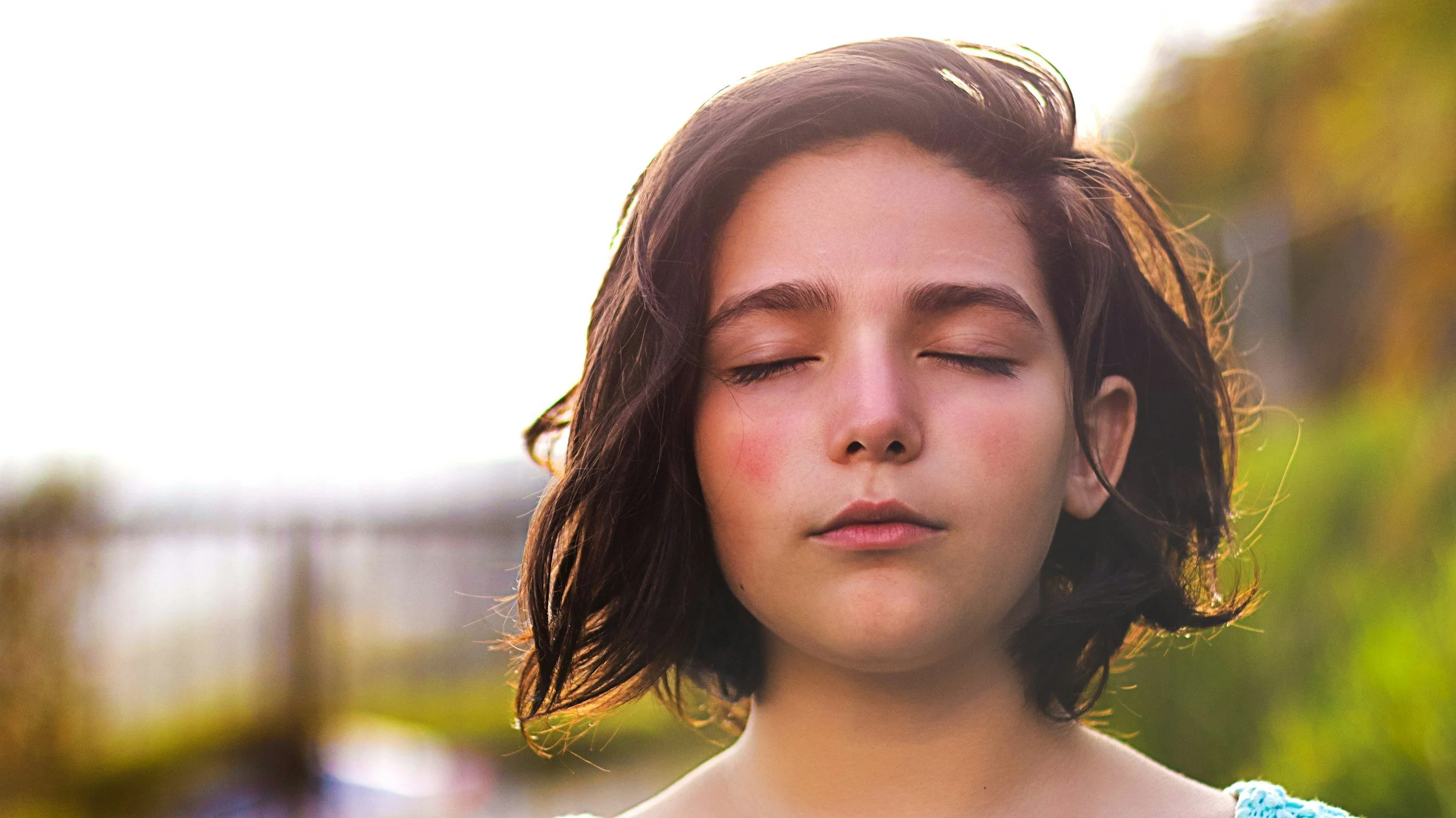 Close-up of a young woman with dark hair and closed eyes, standing outdoors during sunset with a blurred background.
