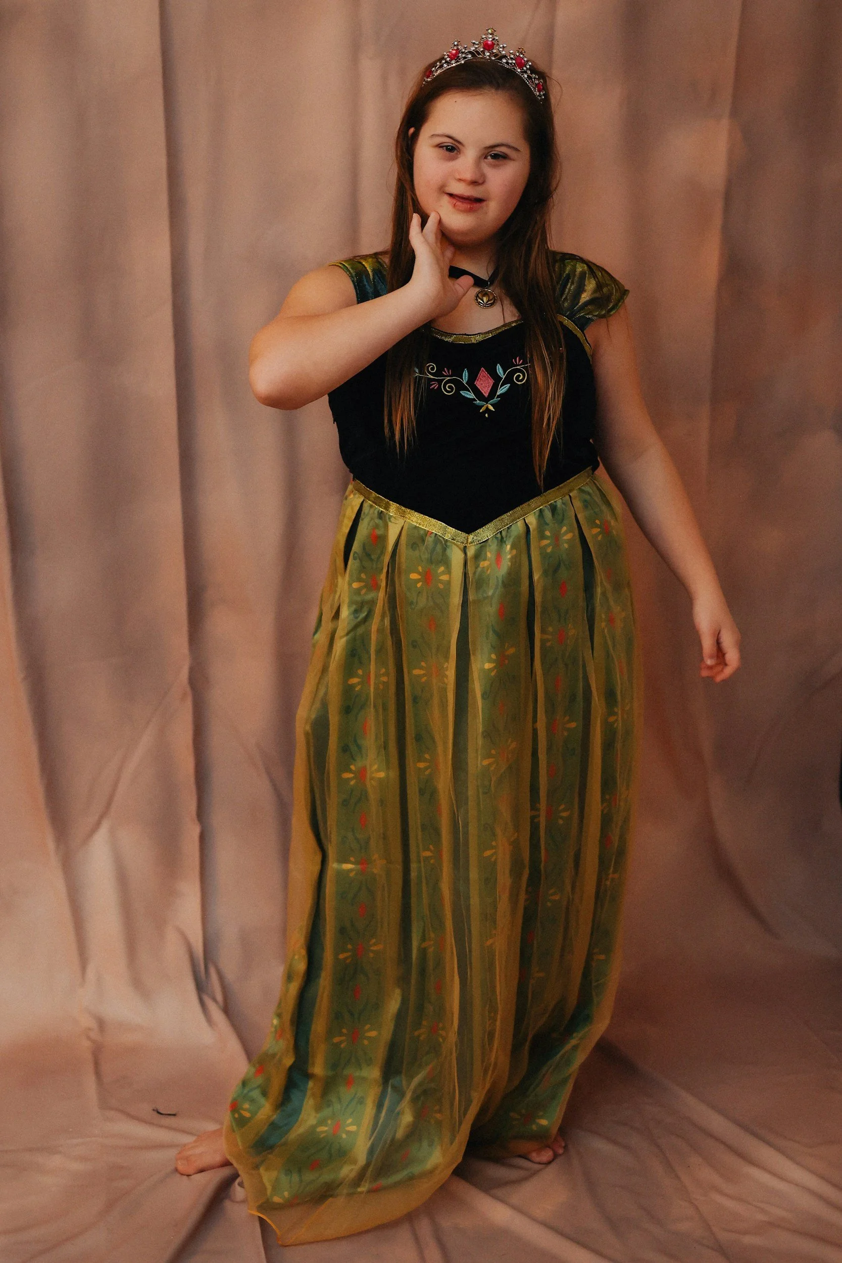 A girl dressed in a princess costume with a crown, standing in front of a beige backdrop, with one hand on her face and a slight smile.
