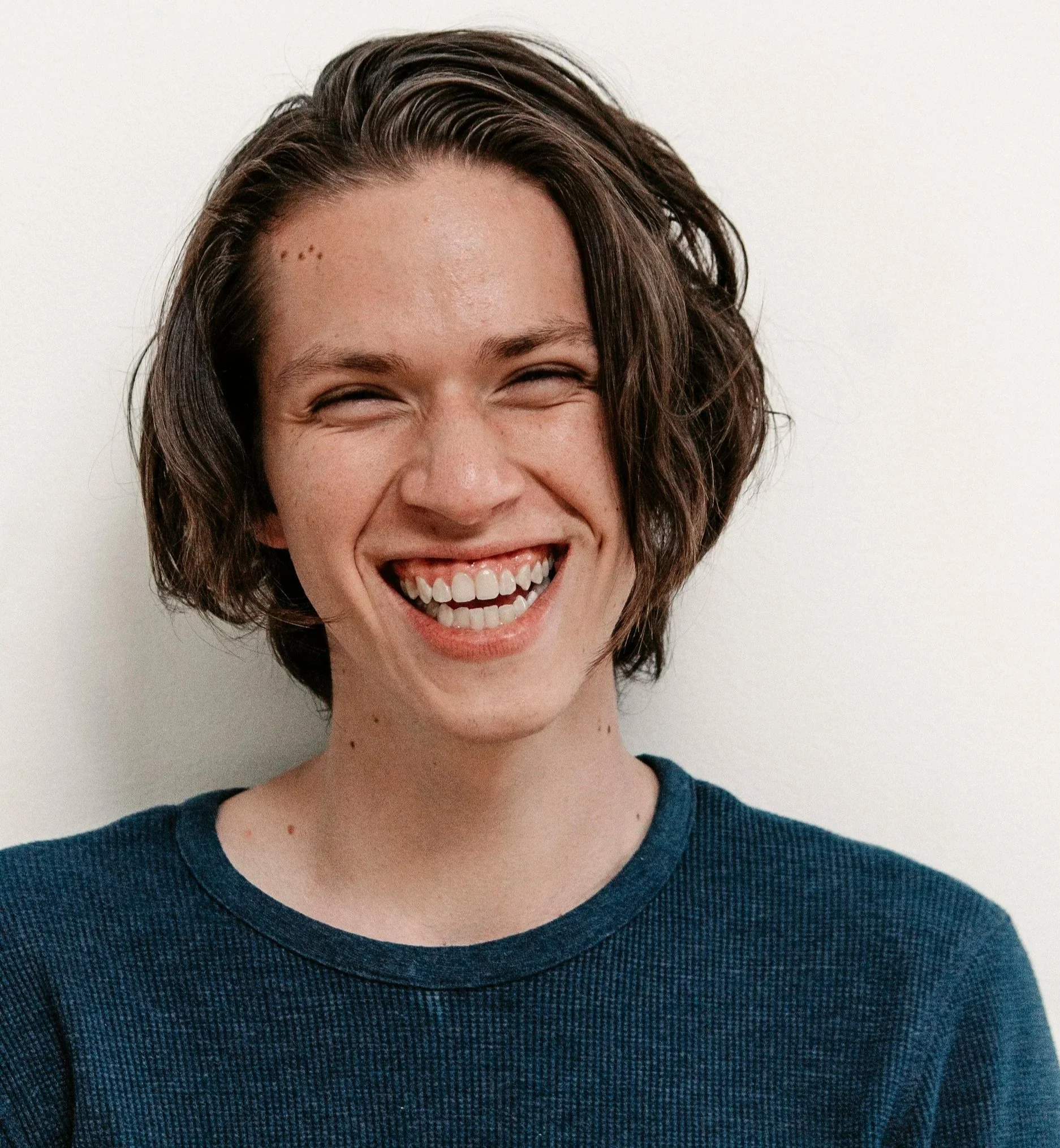 A person with short, wavy brown hair smiling with eyes closed, wearing a dark blue shirt, against a white background.