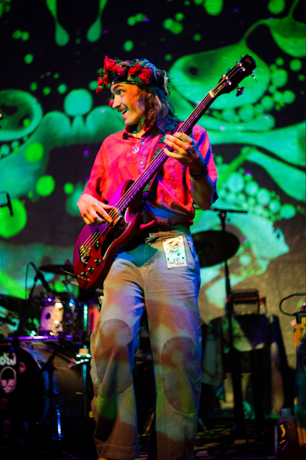 A woman playing a guitar on stage with colorful lights and a green abstract background, wearing a red shirt and a floral crown.