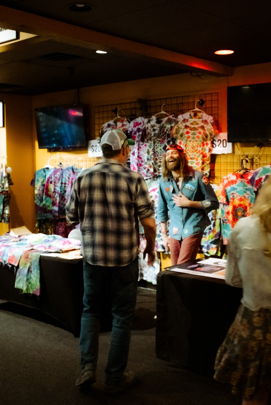 Two men talking at a booth with colorful tie-dye shirts hanging on a wall behind them.