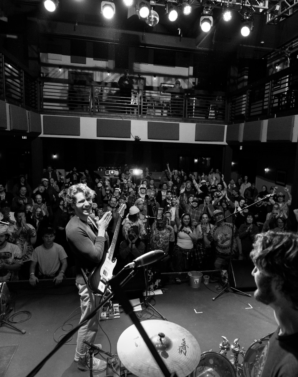 Black and white photo of a band performing on stage in a nightclub with a large audience cheering and clapping.