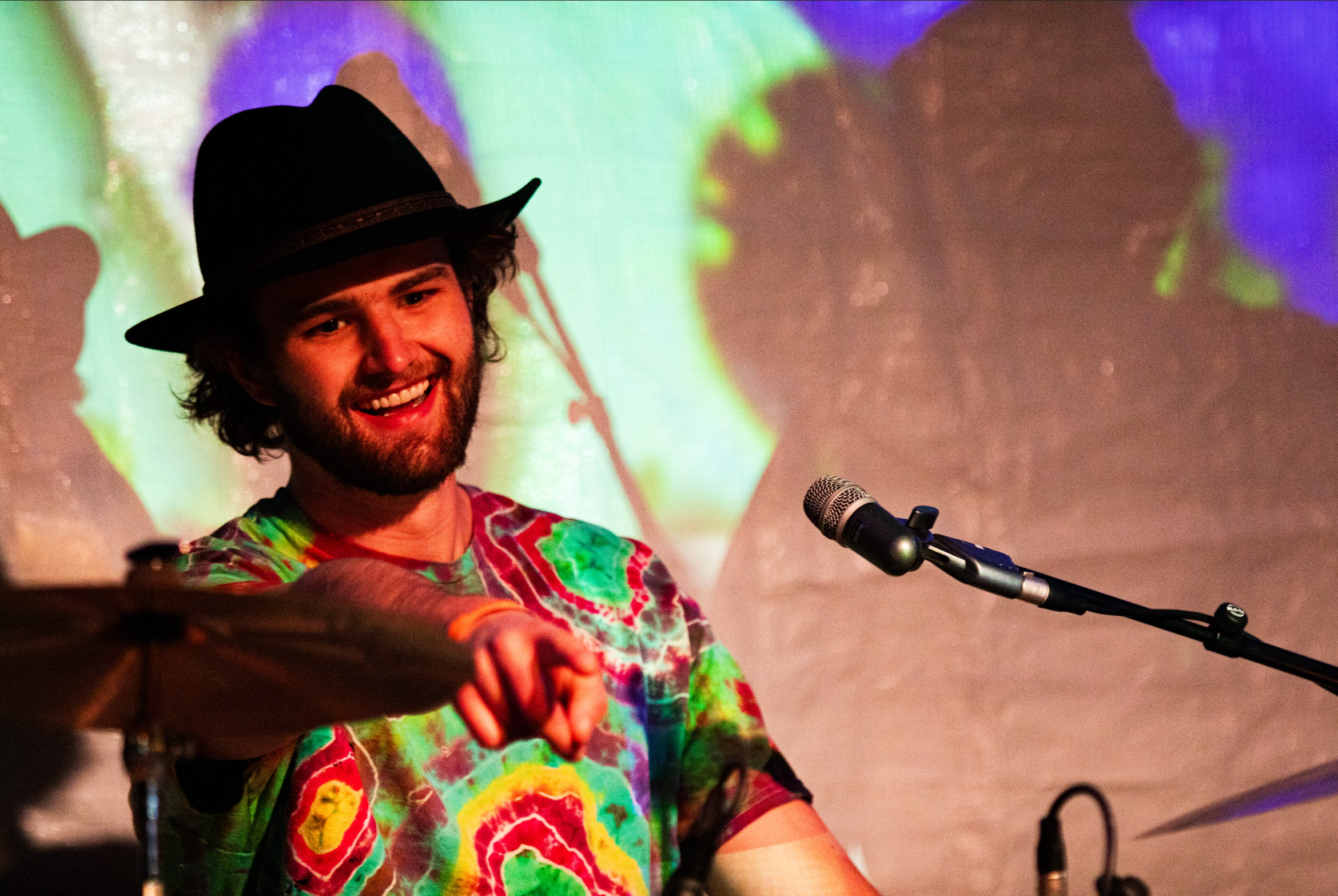 A bearded man with long curly hair wearing a black fedora hat and a colorful tie-dye shirt, smiling and pointing towards the camera with a microphone in front of him, with a vibrant, abstract background.