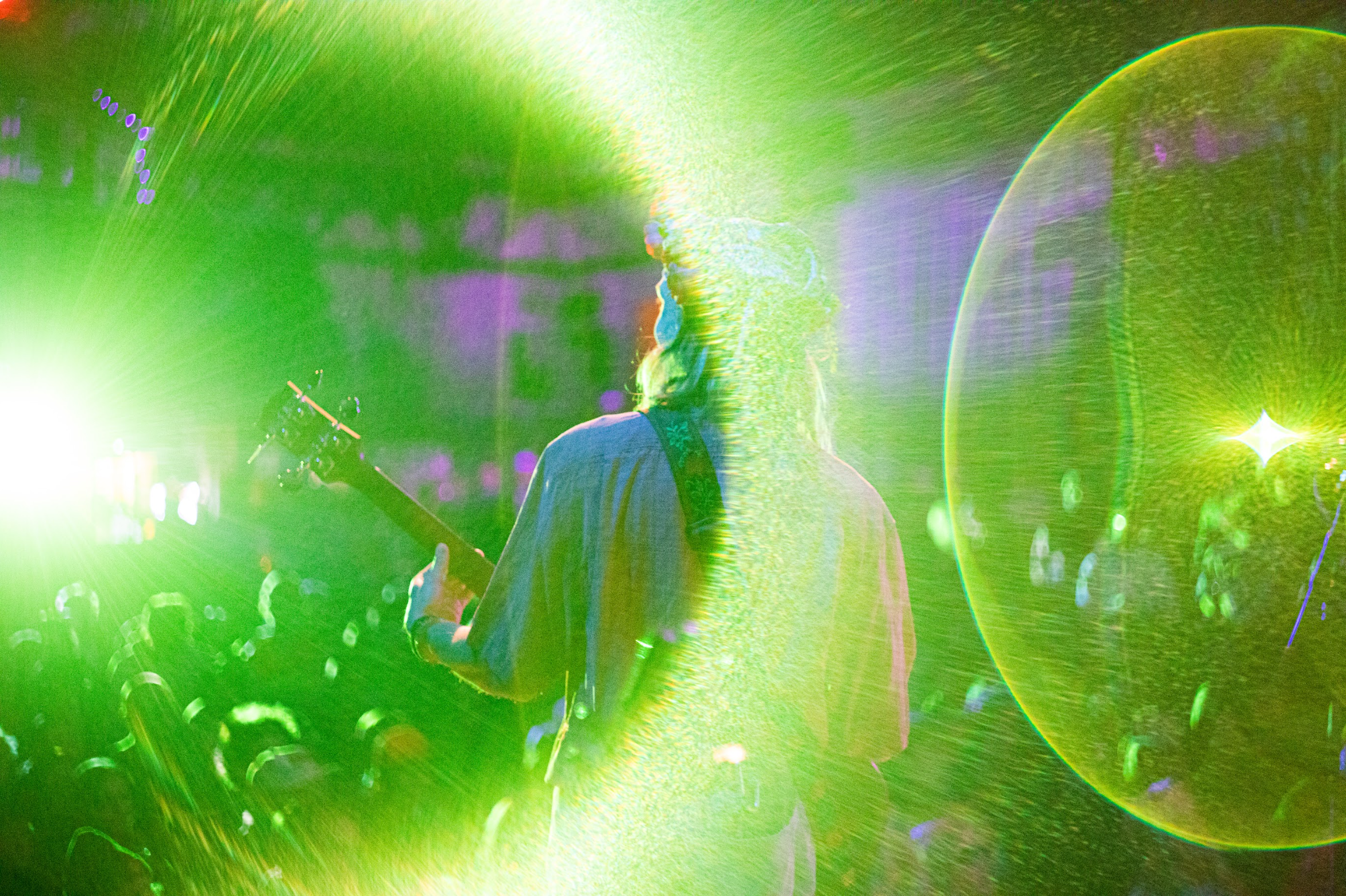 A musician with long hair playing a guitar on stage, with bright green and yellow lights creating a vibrant, energetic atmosphere at a live concert.