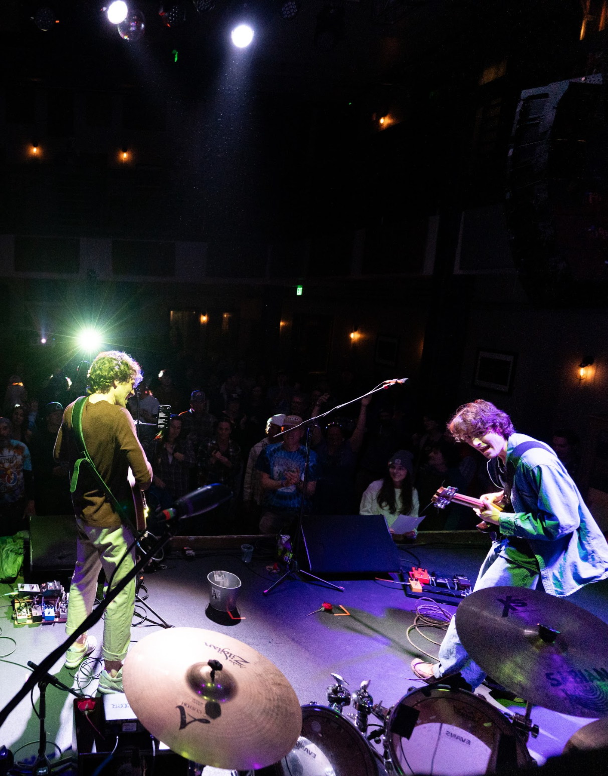 Musicians performing on stage with a band, playing electric guitar and acoustic guitar, while the audience watches in a dimly lit venue with stage lights and musical equipment.