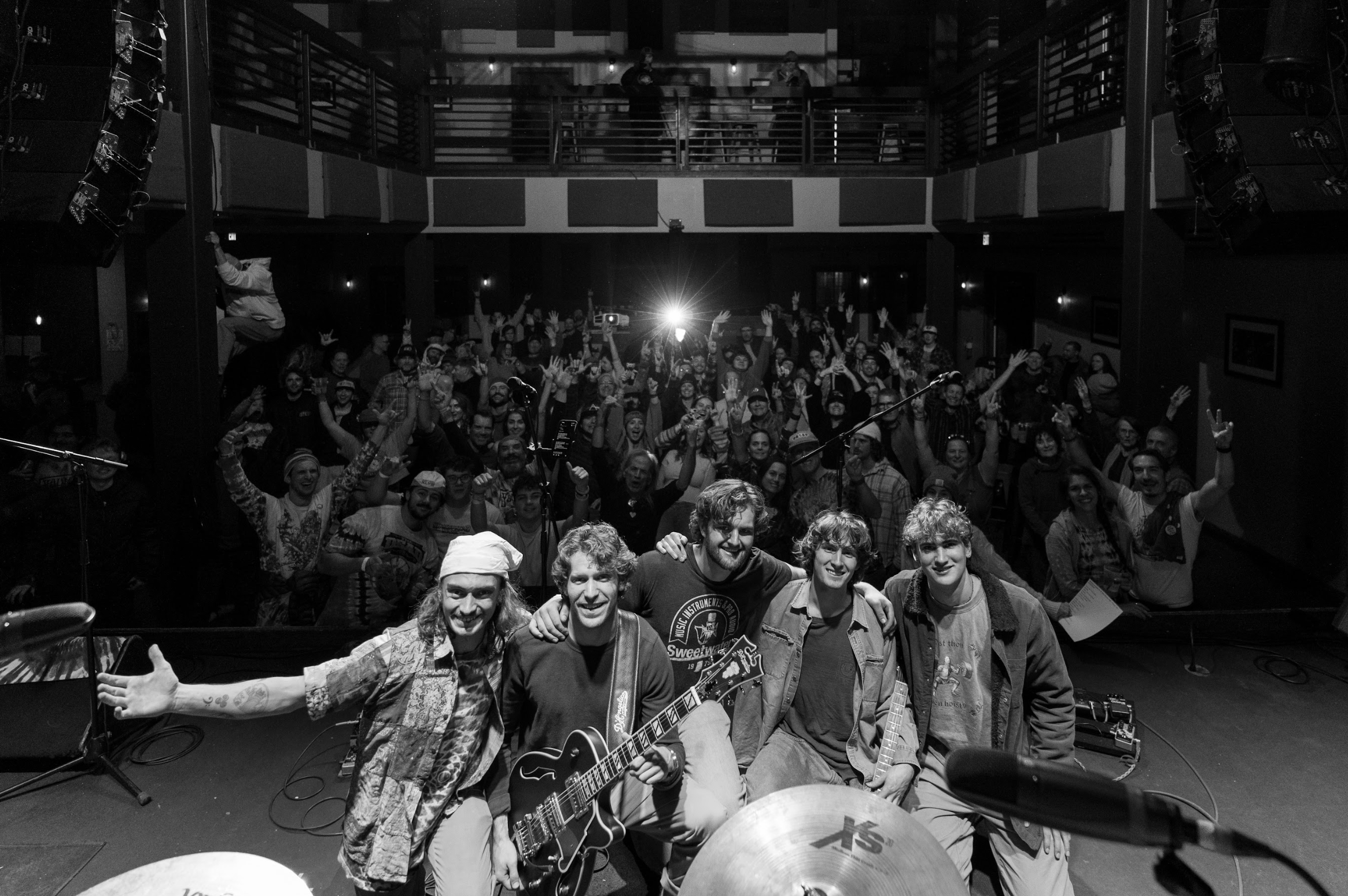 A band takes a bow on stage with a large cheering audience behind them in a concert hall.