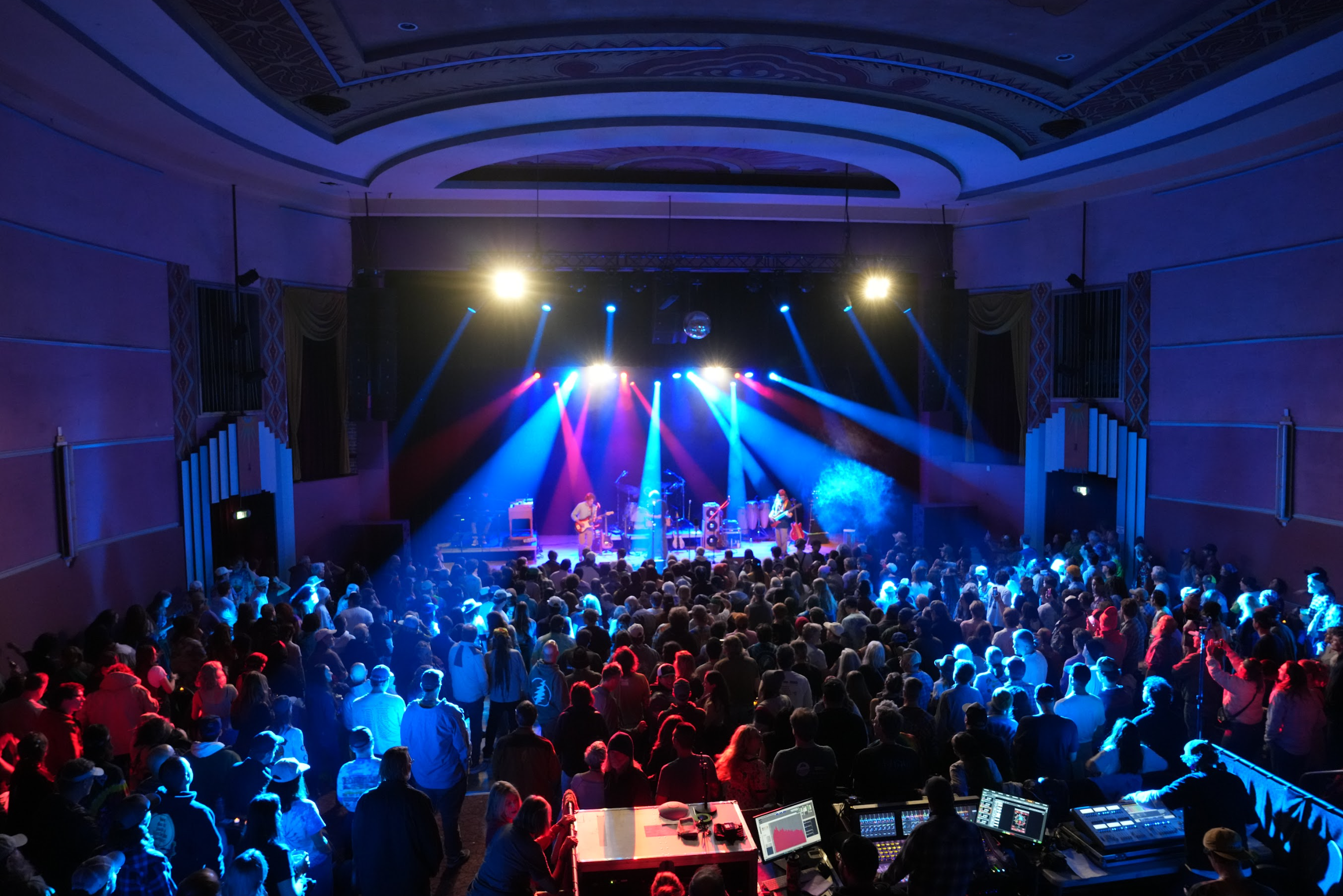 Concert venue with a band performing on stage illuminated by colorful lights, and a large audience watching and enjoying the show.