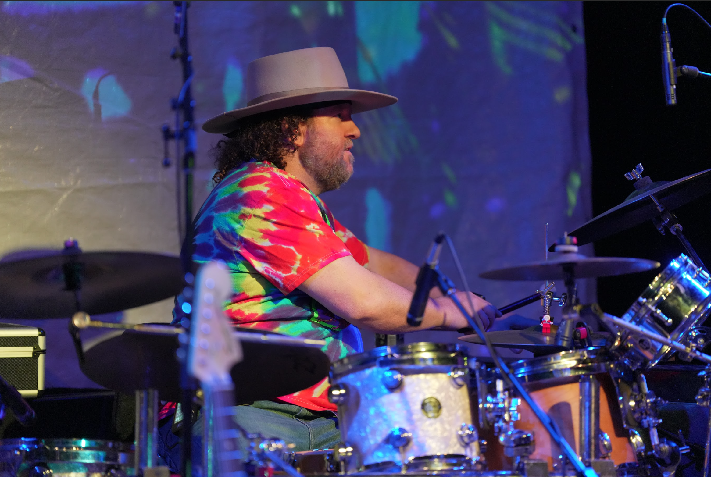 A man wearing a large hat and a colorful tie-dye shirt playing drums on stage, with a backdrop of colorful lights and patterns.