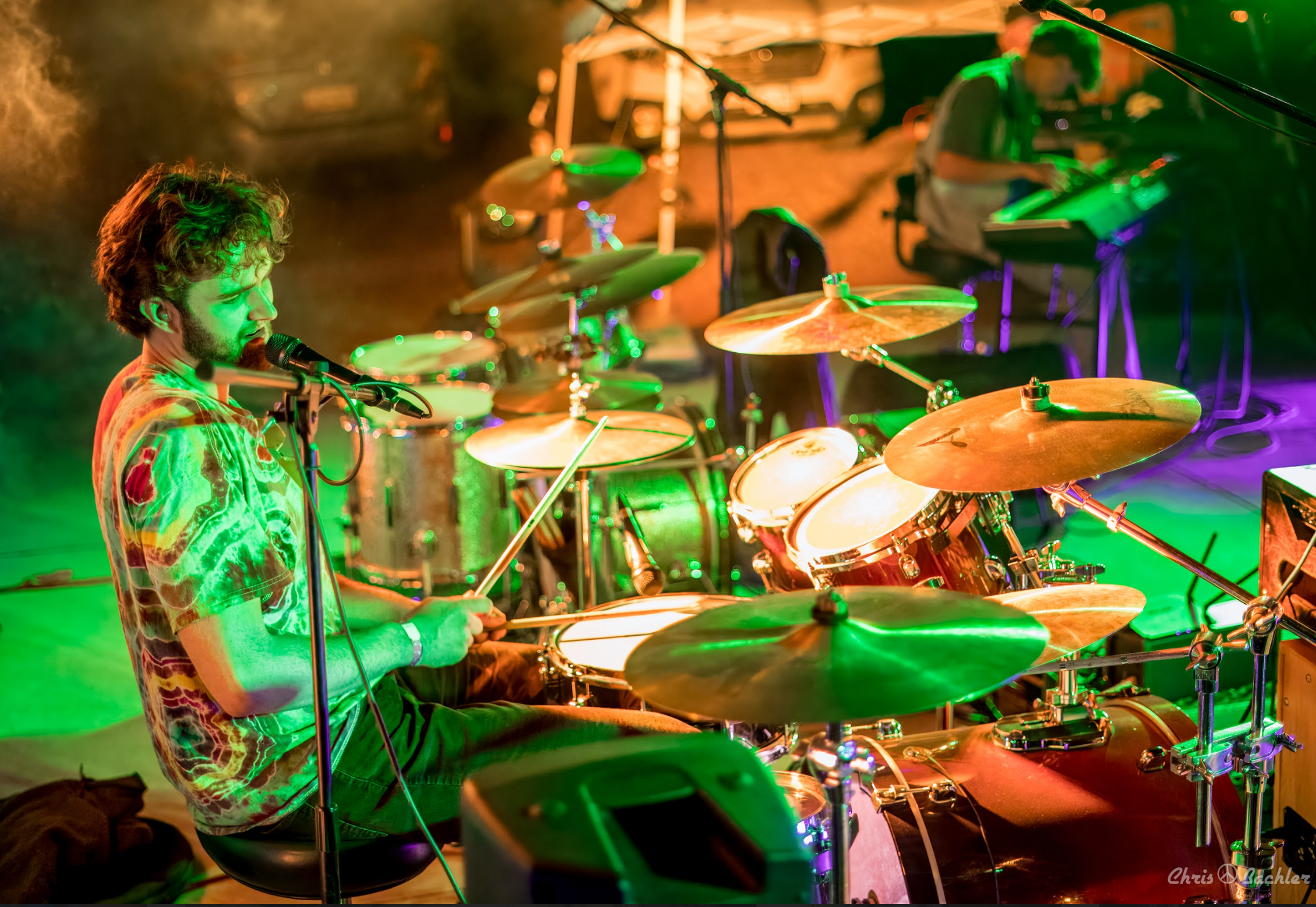 A musician playing drums on stage with colorful green, yellow, and purple lighting, wearing a tie-dye shirt, and singing into a microphone