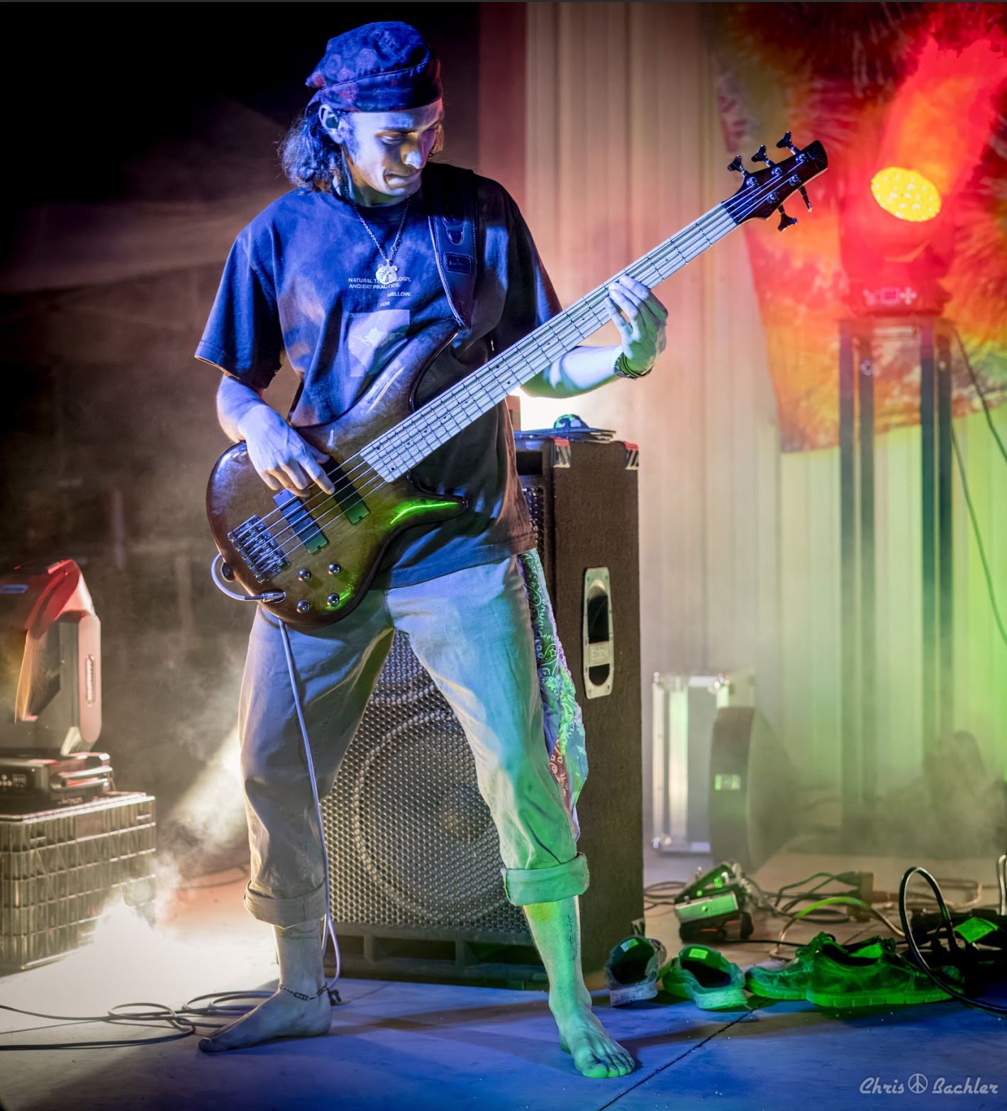 A musician playing a bass guitar on stage, wearing a dark t-shirt, light-colored rolled-up pants, barefoot, with long hair and a headband. Stage lights and equipment are visible around him.