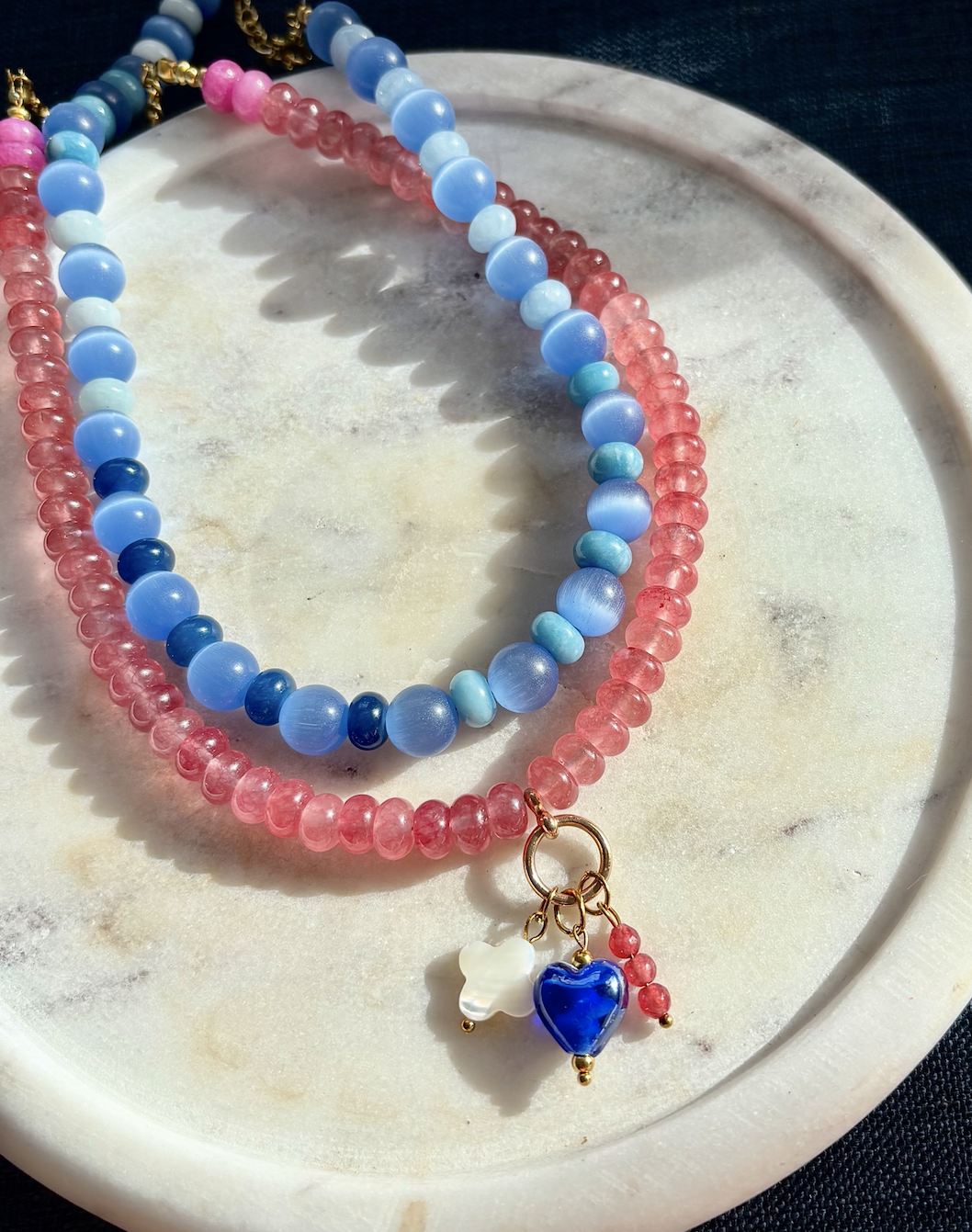 Two beaded necklaces, one with pink beads and the other with blue beads, displaying a dangling blue heart and white flower charm on a white circular surface.