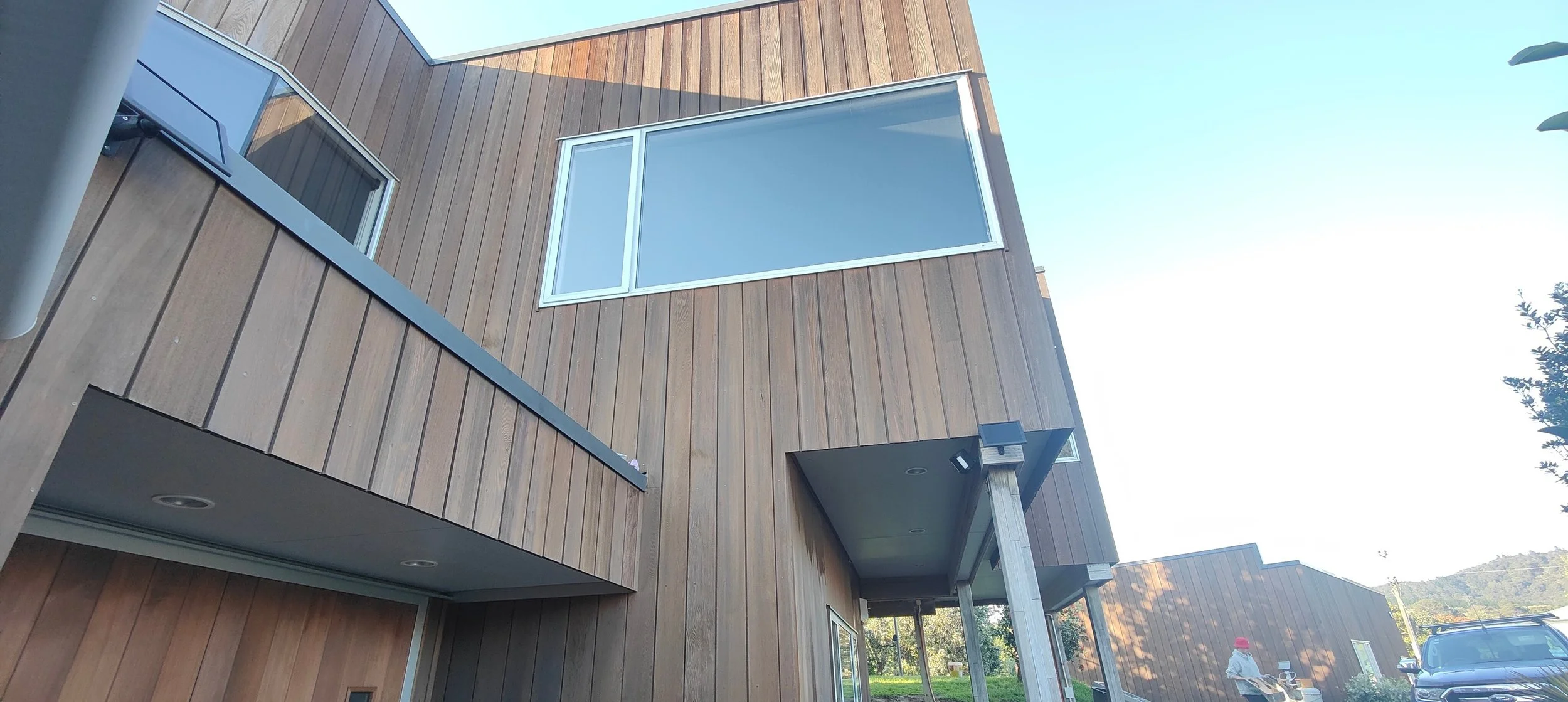Modern multi-story building with wood siding, large windows, and an outdoor balcony, with a person in a red hat and a vehicle visible in the background.