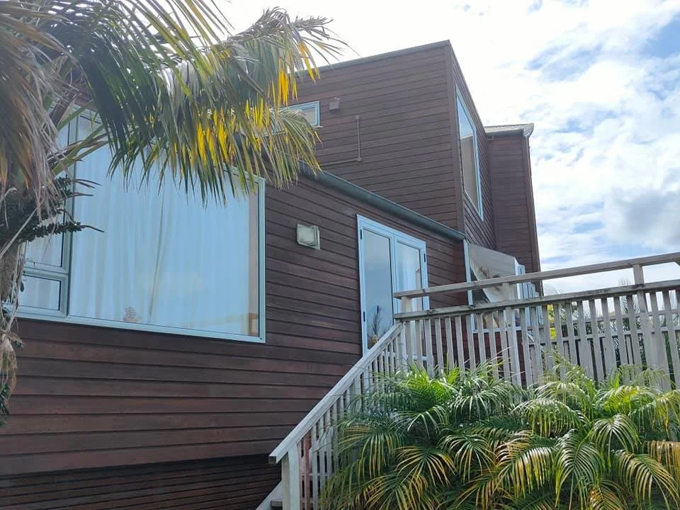 A two-story house with brown siding, large windows, a white railing porch, and lush green plants in front.