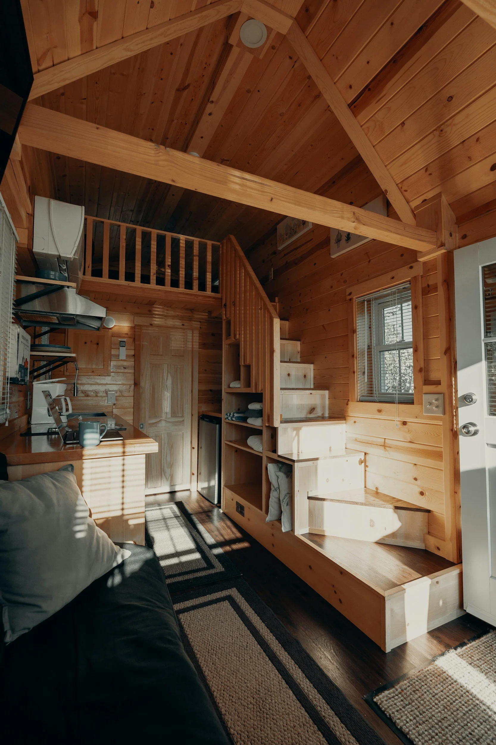 Interior of a cozy wood cabin with a loft, staircase, small kitchen area, and sunlight streaming through windows.