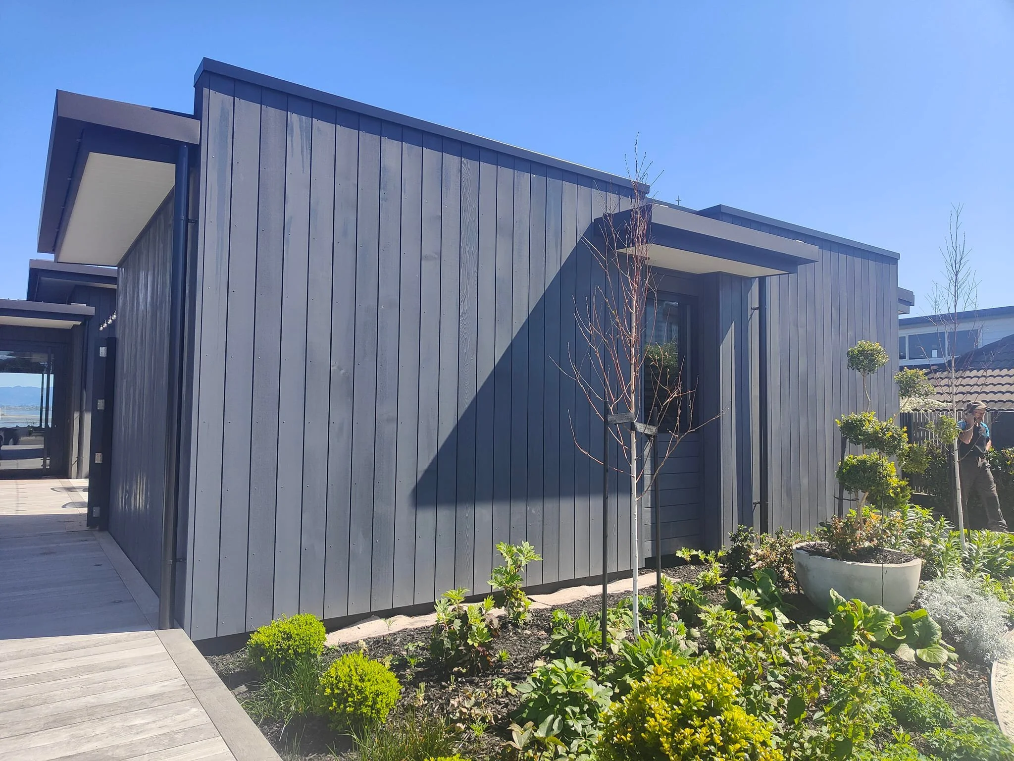 Modern house with dark gray wooden exterior, small garden with various plants, and a bright blue sky.