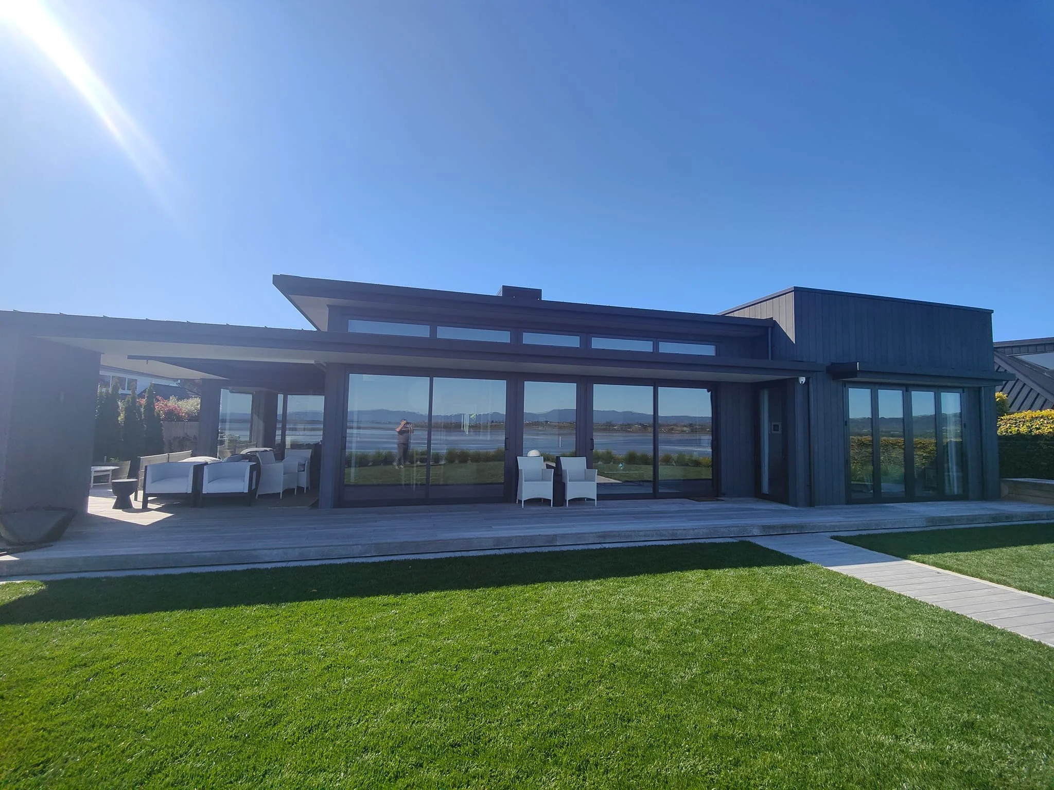 Modern house with large glass windows and patio furniture on wooden deck, green lawn, clear blue sky, and water view in the background.