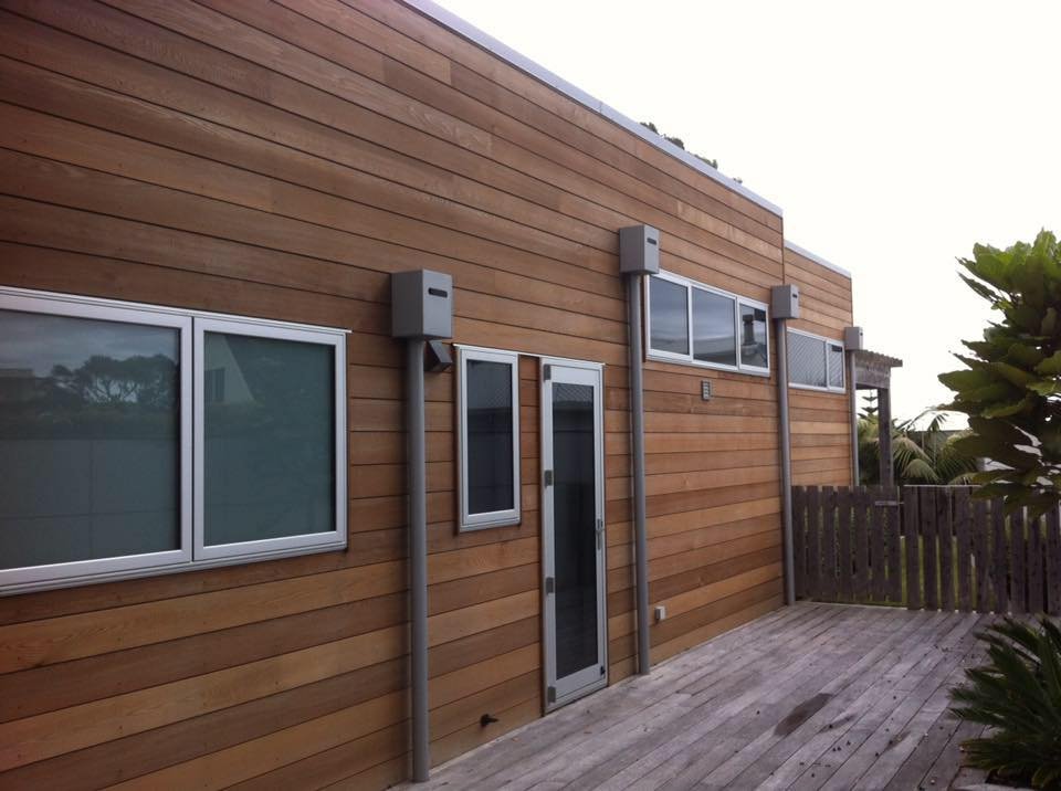 Back patio of a modern house with horizontal wooden siding, several windows, and a glass door, enclosed by a wooden fence.