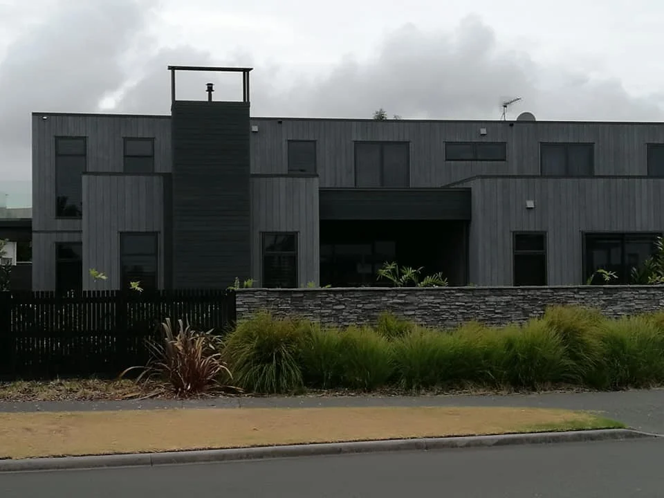 Modern gray building with multiple windows, surrounded by a landscaped garden with green plants and a stone wall in front.