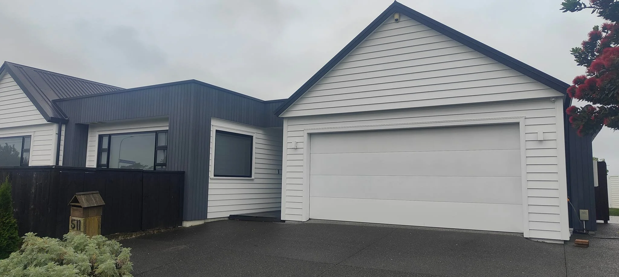 Modern house with white and dark gray siding, black fence, large garage door, and surrounding plants, overcast sky.