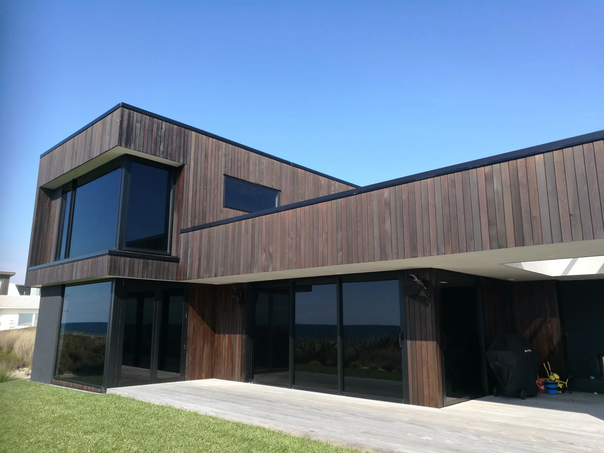 Modern two-story house with wooden exterior, large glass windows, and a deck, near the beach under a clear blue sky.