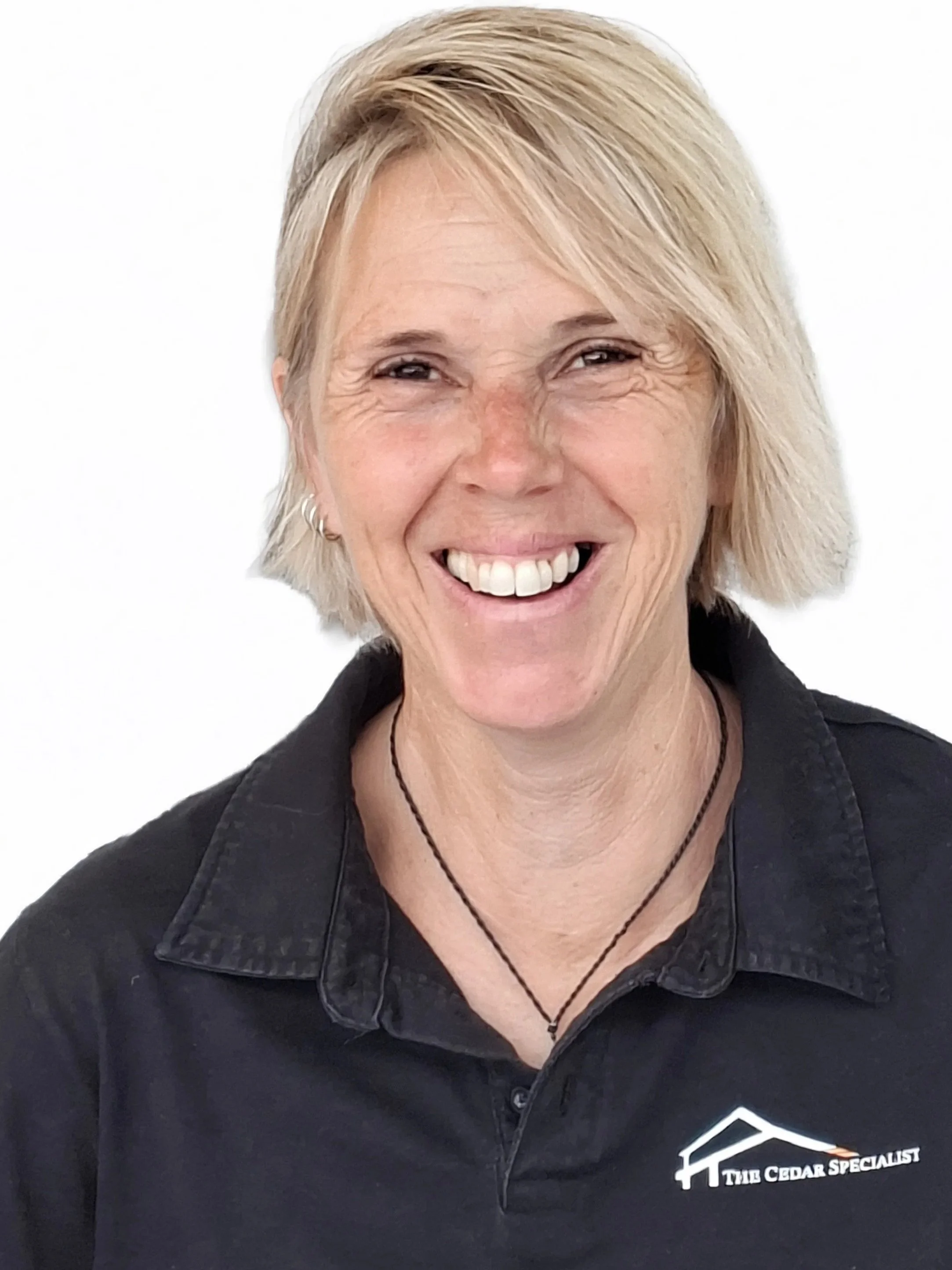 A woman with blonde hair smiling, wearing a black polo shirt with a logo that says 'The Cedar Specialist' and a necklace, standing against a white background.