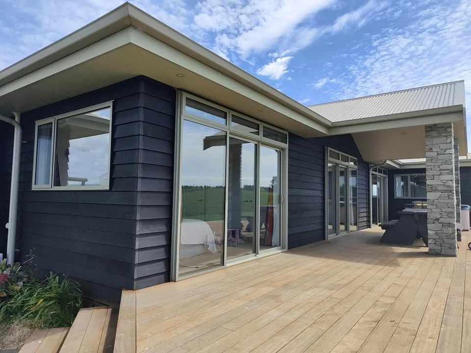 Modern house with dark blue exterior siding, large glass sliding doors, and a wooden deck, under a partly cloudy sky.