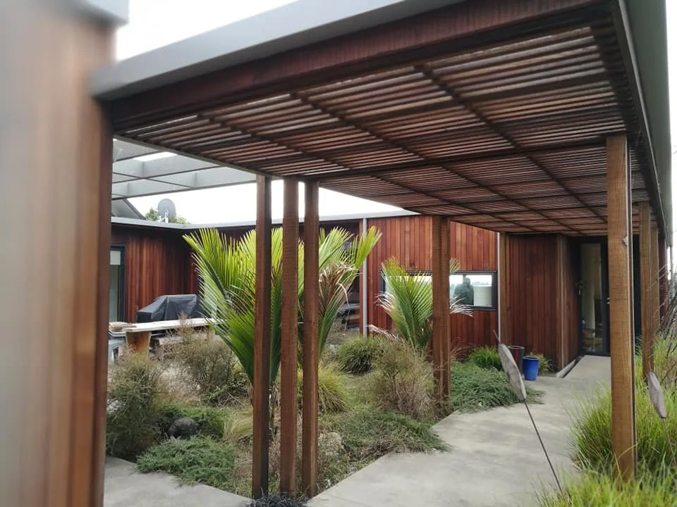 A covered outdoor patio with a wood slat roof supported by wooden posts, surrounded by desert-style plants and shrubs, with a concrete pathway and a building with wooden siding in the background.