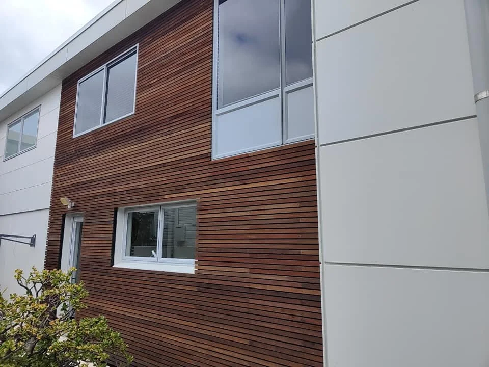 Modern residential building exterior with white panels and wood accent siding, featuring multiple windows and a small tree in the foreground.