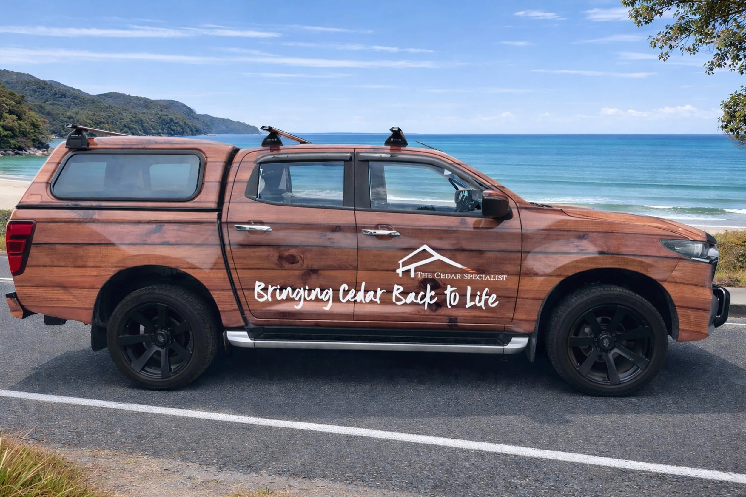 A pickup truck with a wood grain exterior parked by a beach, with ocean and mountains in the background. The truck has a logo and text that reads 'The Cedar Specialist' and 'Bringing Cedar Back to Life'.