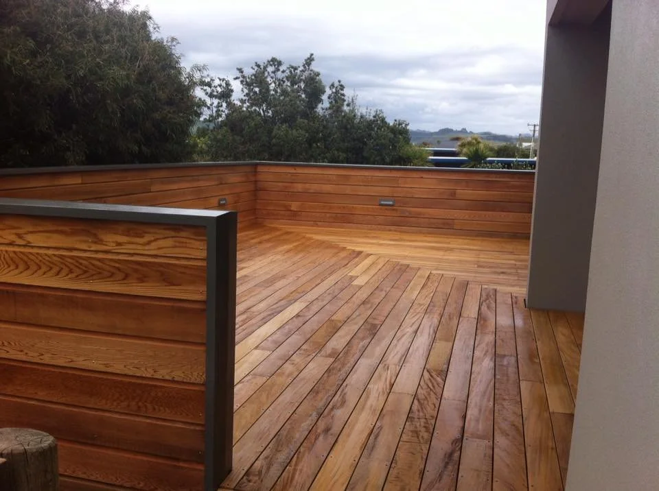 Empty wooden deck with a low wooden railing and gray wall, overlooking trees and cloudy skies in the distance.